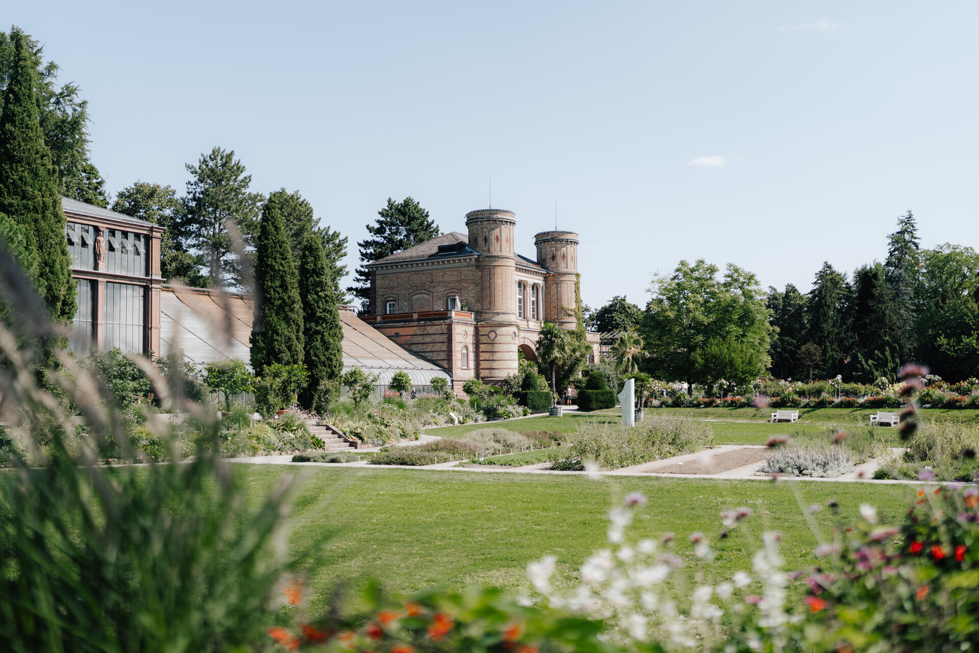 Ein historisches Backsteingebäude mit zwei runden Türmen steht im Hintergrund, umgeben von üppigen Gärten und gepflegten Rasenflächen - eine idyllische Kulisse für eine Hochzeit bei Tante Polly in Karlsruhe unter strahlend blauem Himmel.