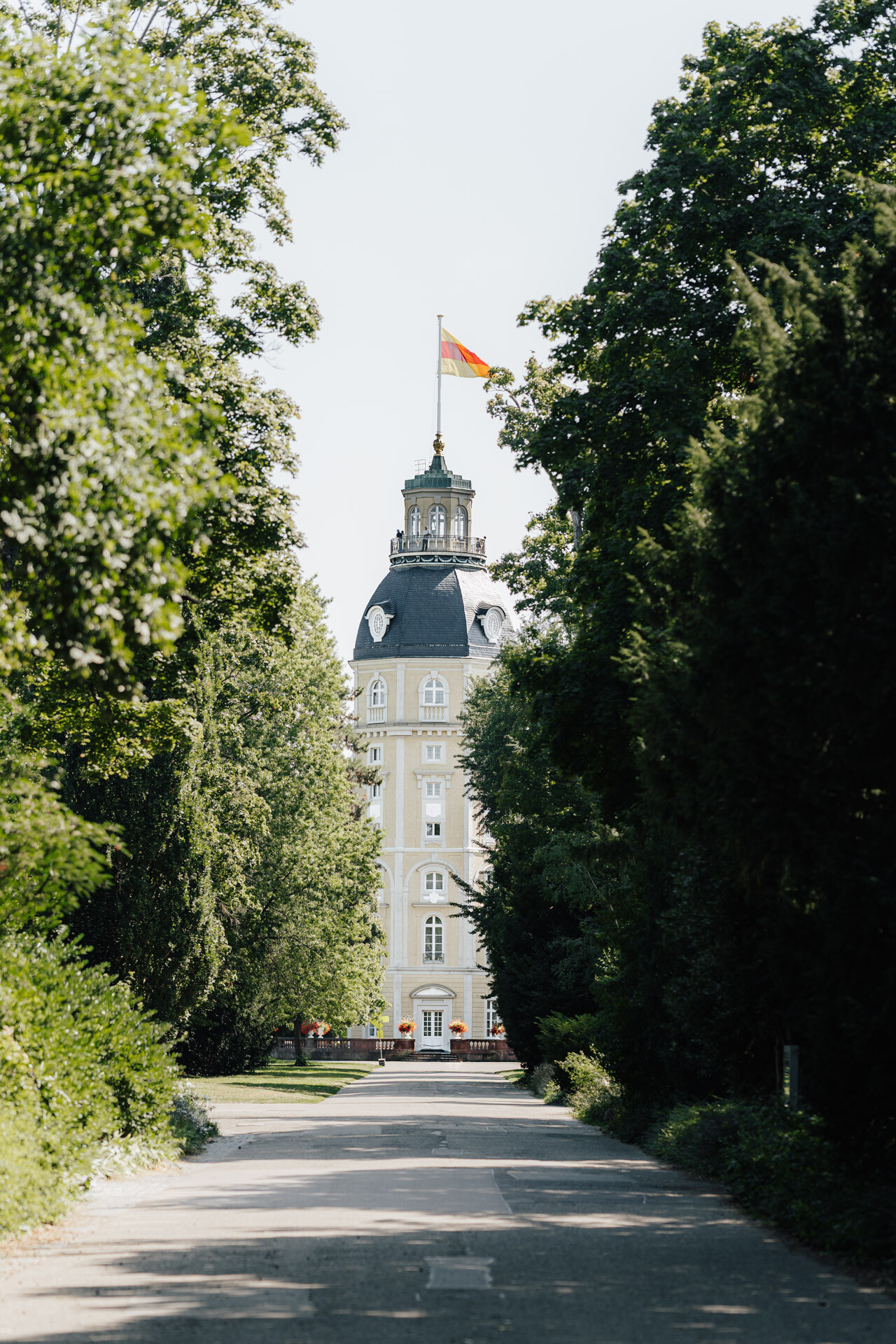 Ein hohes, historisches Gebäude mit einem Kuppeldach und einer Fahne auf der Spitze steht am Ende eines von Bäumen gesäumten Weges an einem sonnigen Tag in Karlsruhe, perfekt für eine Hochzeit oder ein Treffen bei Tante Polly.