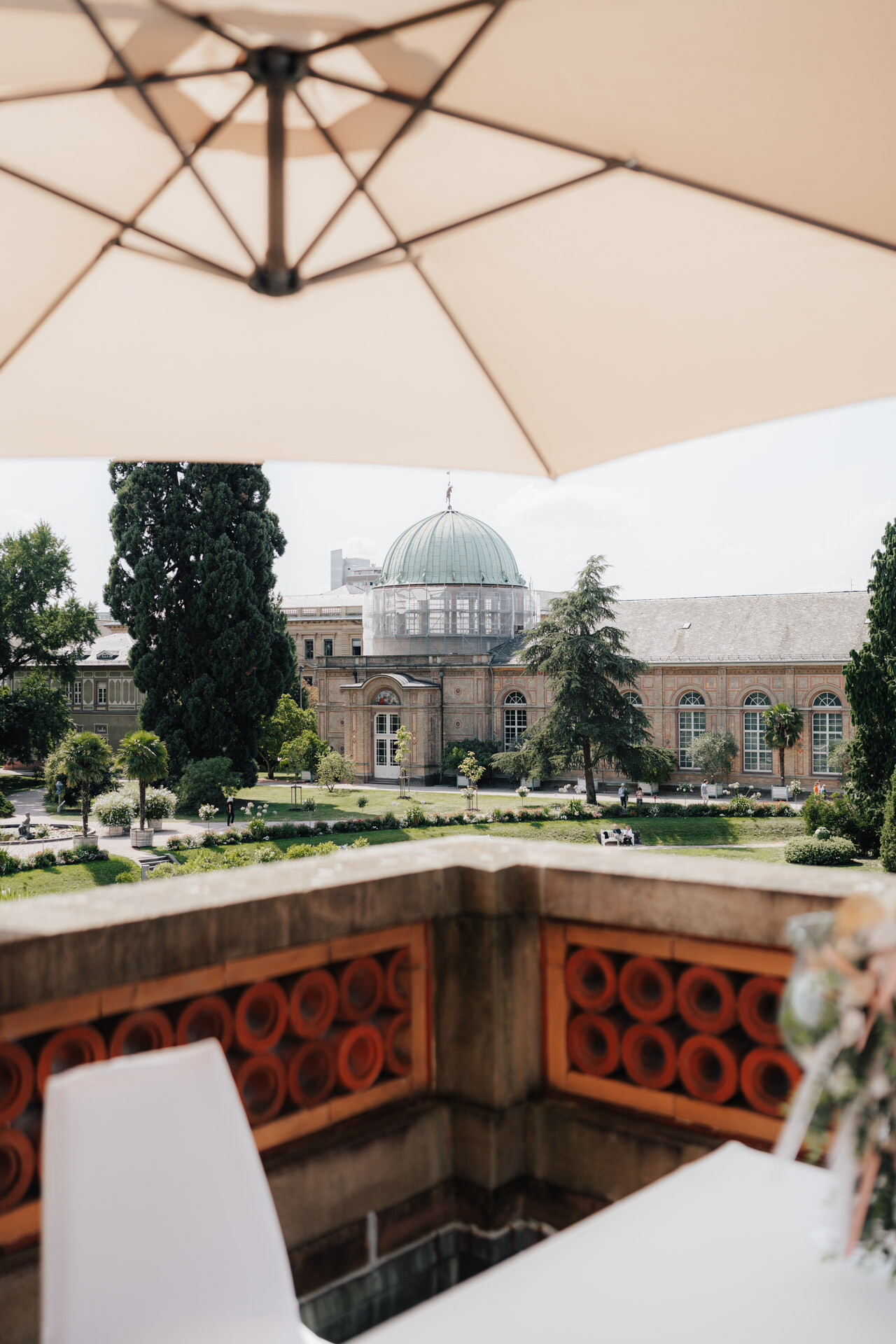 Blick unter einem großen Terrassenschirm auf Hochzeit Tante Polly Karlsruhe, mit Blick auf ein historisches Gebäude mit einem gewölbten Glasdach und Bogenfenstern, umgeben von grünen Bäumen und Gärten. Ein weißer Stuhl und ein Blumenarrangement zieren den Vordergrund.