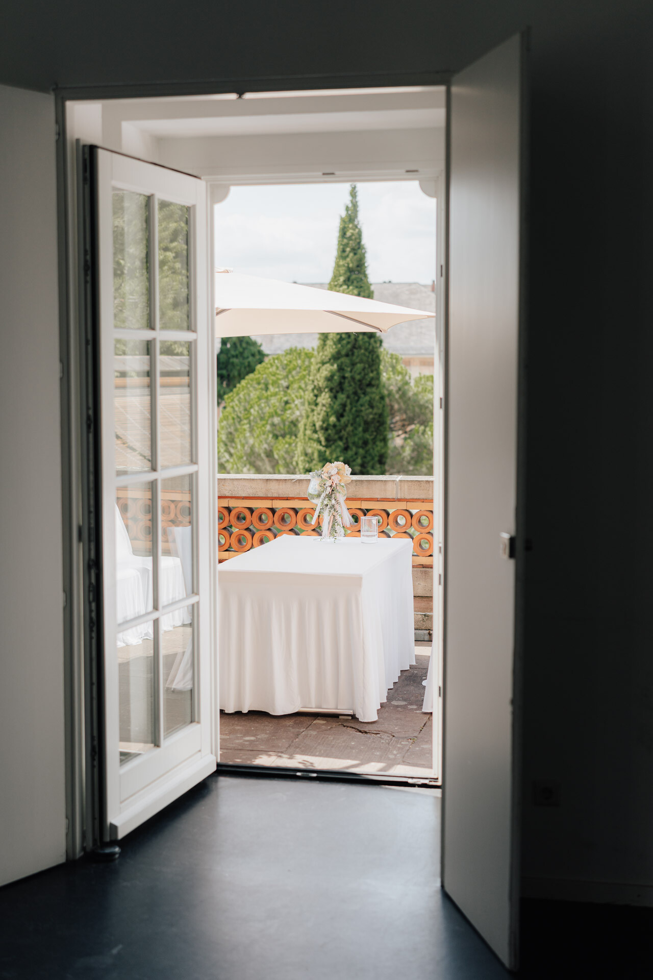 Eine Glastür öffnet sich zu einem Balkon der Hochzeit Tante Polly Karlsruhe, auf dem ein mit einem weißen Tuch bedeckter Tisch mit einer Vase voller Blumen steht. Draußen sind ein Sonnenschirm und üppiges Grün zu sehen, durch das natürliches Licht fällt.