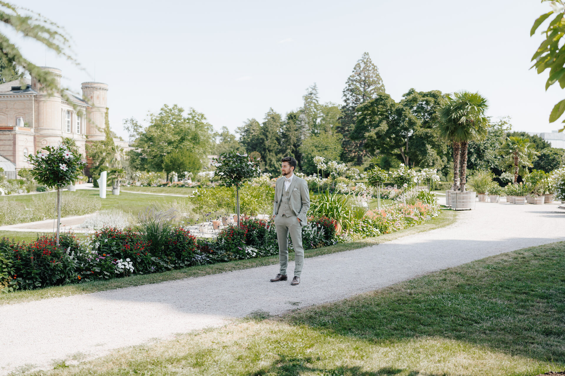 Ein Mann in einem hellgrauen Anzug steht auf einem Kiesweg in einem Landschaftsgarten der Hochzeit Tante Polly Karlsruhe, in dem bunte Blumenbeete, grüner Rasen, Bäume und ein historisches Gebäude unter einem klaren Himmel zu sehen sind.