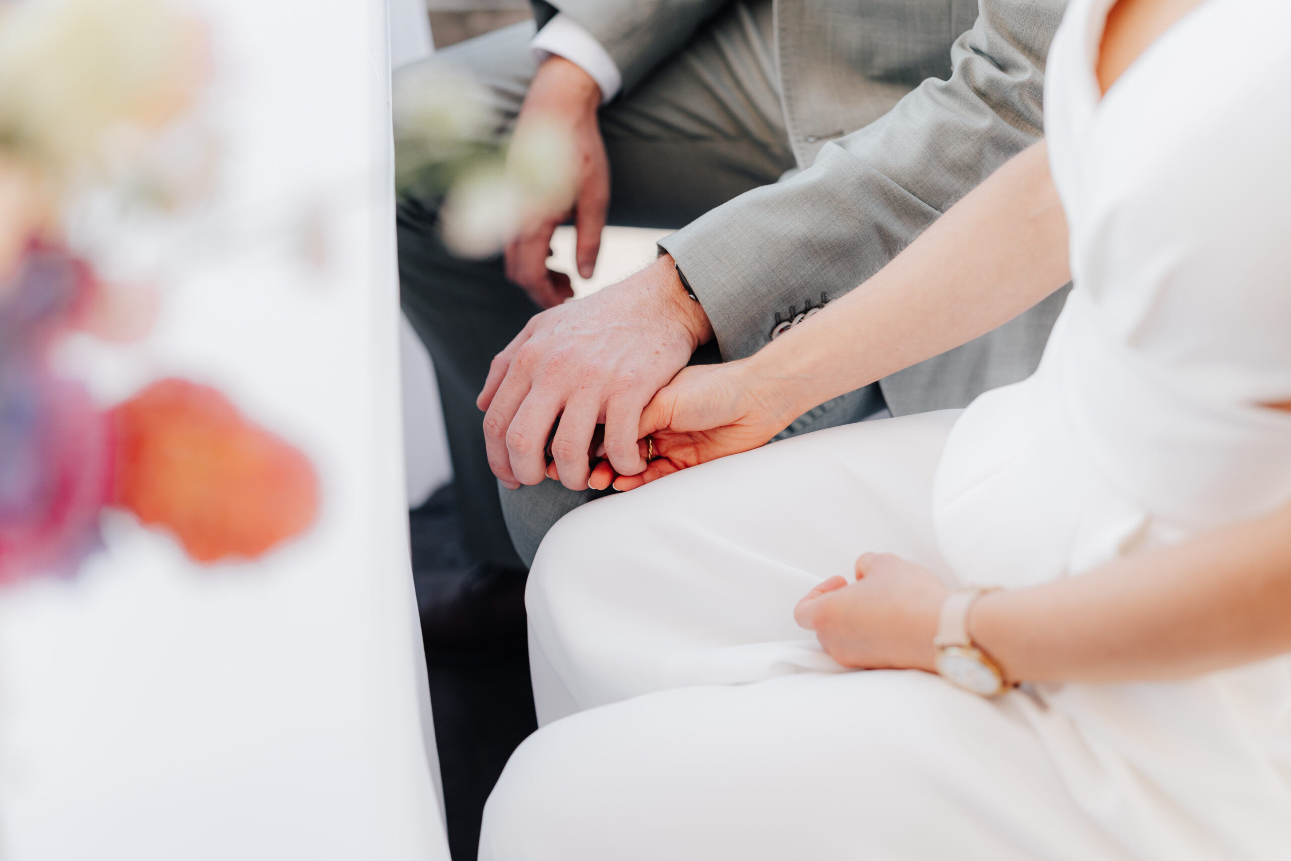 Nahaufnahme von Braut und Bräutigam, die sich bei ihrer Hochzeit in Karlsruhe an den Händen halten. Die Braut trägt ein weißes Kleid und eine Uhr, während der Bräutigam einen hellgrauen Anzug trägt - ihre Hände sind in einem intimen, zärtlichen Moment bei Tante Polly sanft umschlungen.