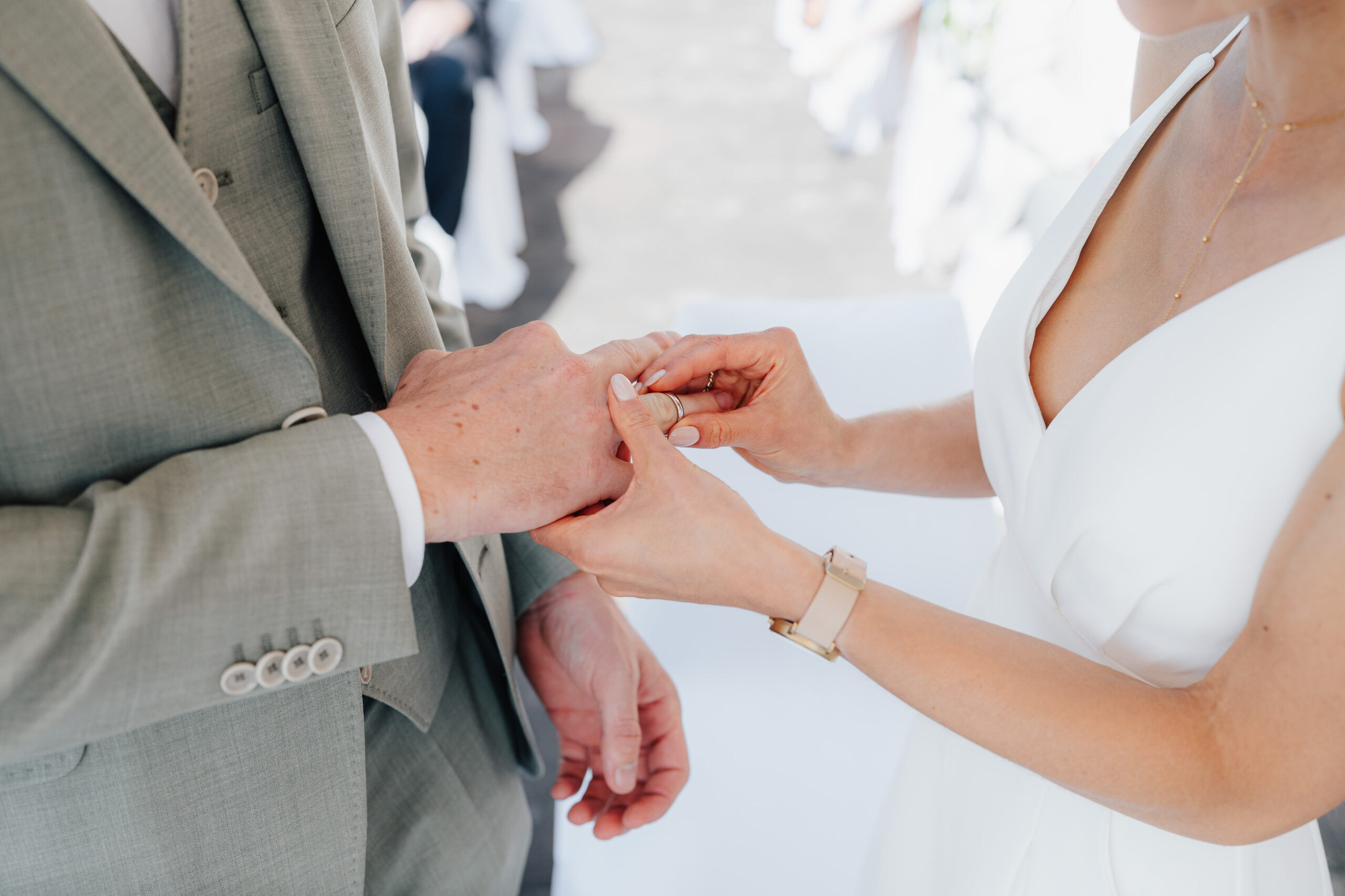 Nahaufnahme einer Braut in einem weißen Kleid, die dem Bräutigam während einer Hochzeit bei Tante Polly Karlsruhe einen Ring an den Finger steckt. Der Bräutigam, in einem hellgrauen Anzug, steht dicht daneben, während sich der Moment auf ihre Hände konzentriert.