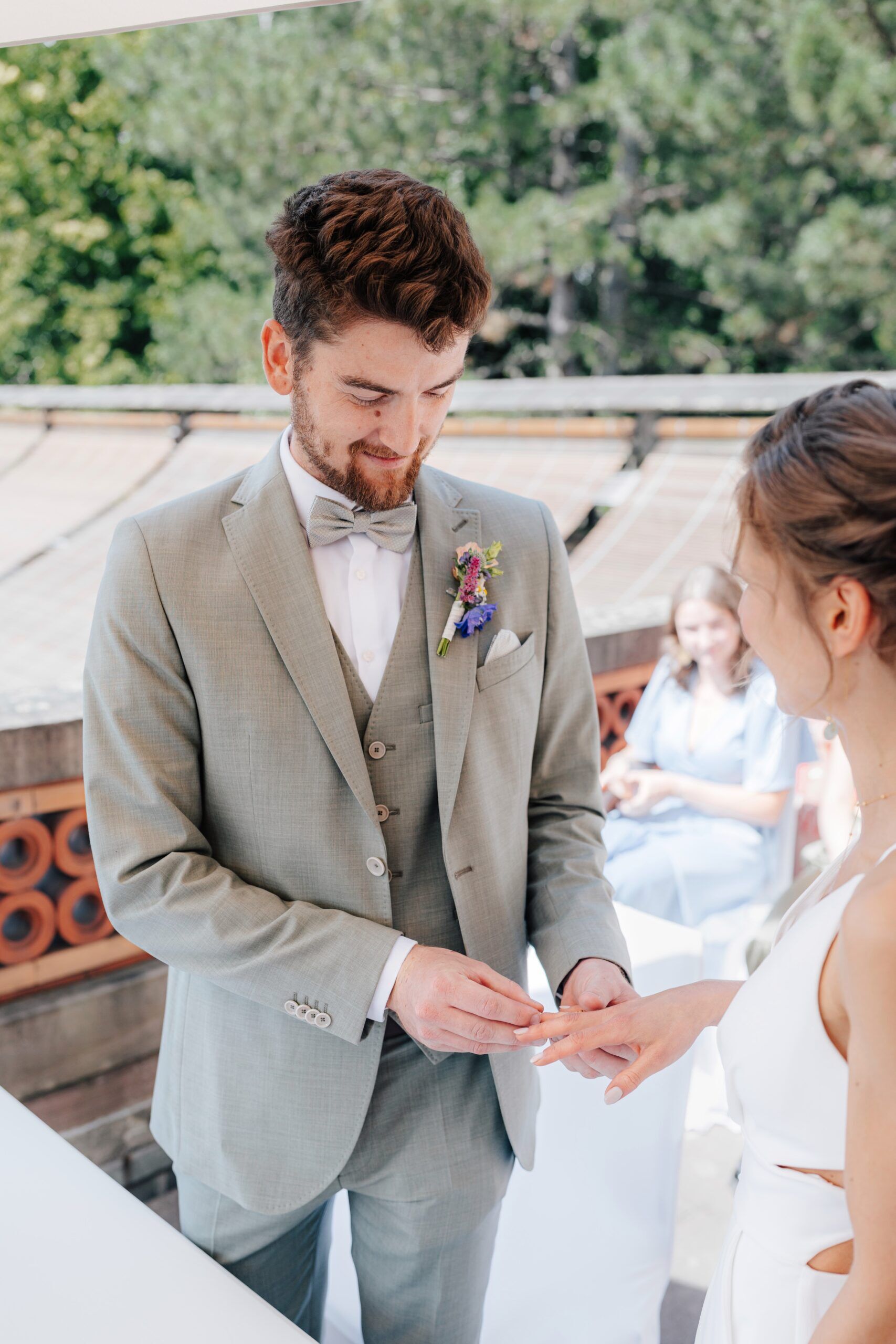 Ein Bräutigam in einem hellgrauen Anzug steckt der Braut während einer Hochzeit im Freien bei Tante Polly Karlsruhe einen Ring an den Finger, mit üppigem Grün und einem sitzenden Gast im Hintergrund.