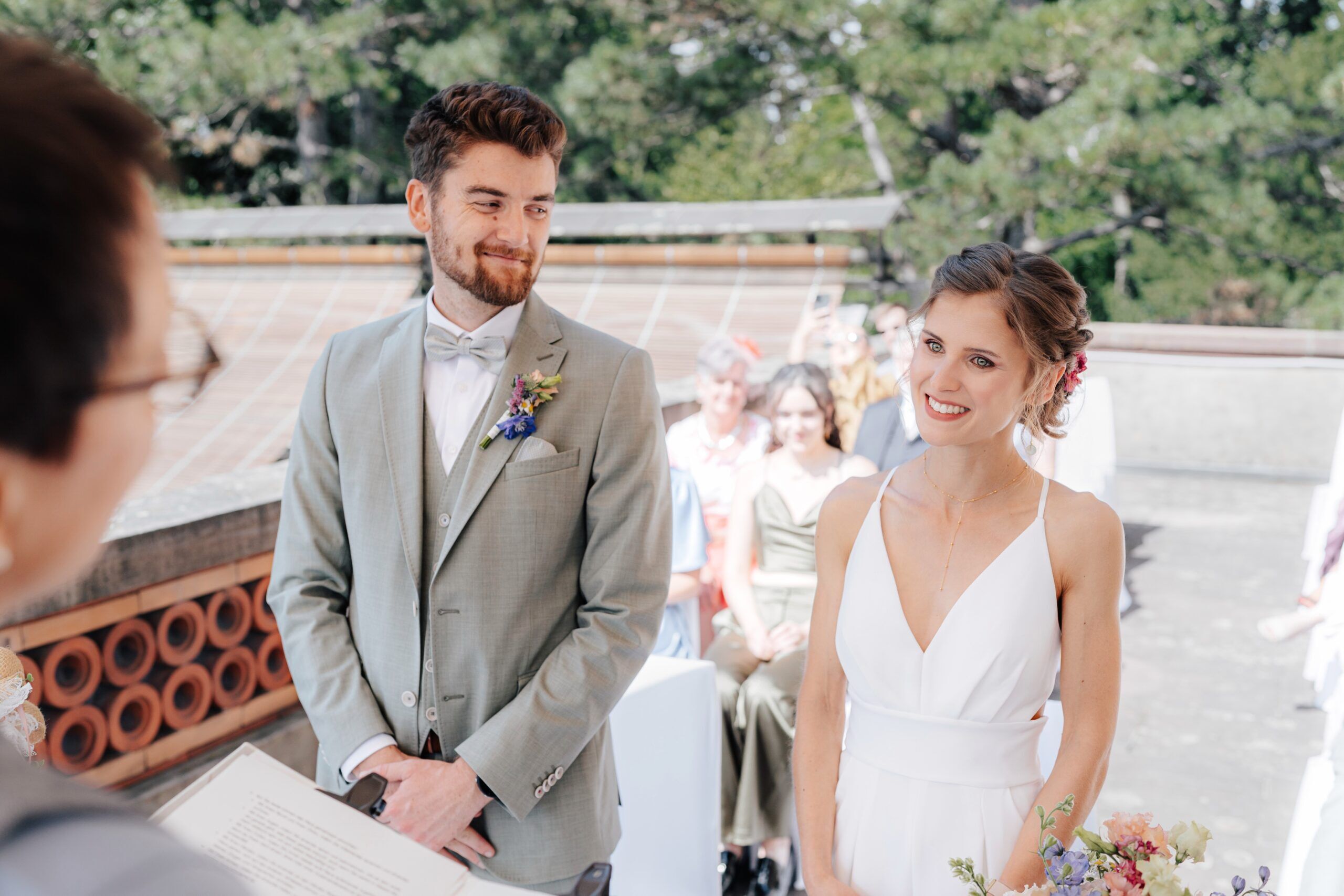Eine Braut in einem weißen Kleid und ein Bräutigam in einem hellgrauen Anzug stehen Seite an Seite im Freien bei Hochzeit Tante Polly Karlsruhe und lächeln dem Trauredner zu, während die Gäste an einem sonnigen Tag im Hintergrund sitzen.