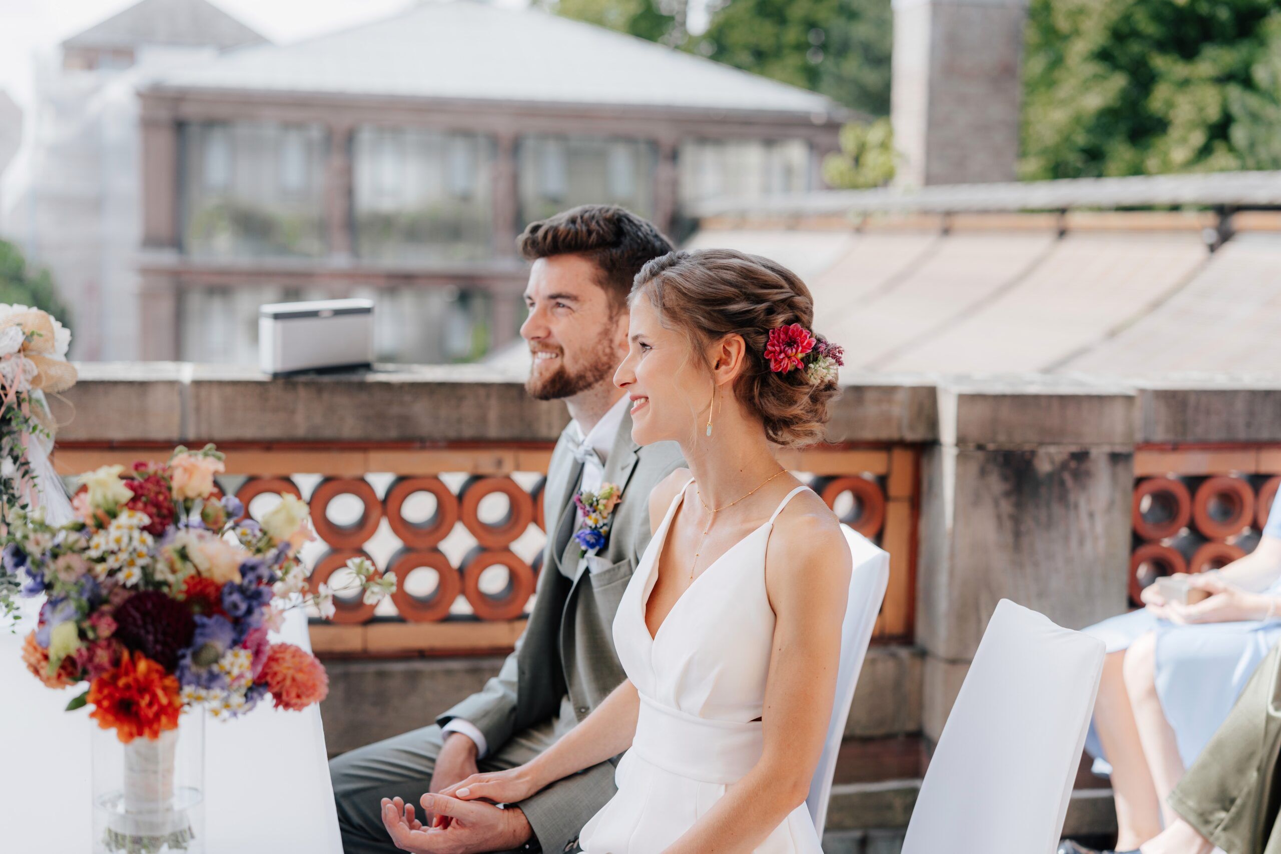 Braut und Bräutigam sitzen Seite an Seite und lächeln während ihrer Hochzeit im Freien bei Tante Polly Karlsruhe. Die Braut trägt ein weißes Kleid mit Blumen im Haar, während der Bräutigam einen hellen Anzug trägt. Bunte Blumen schmücken einen Tisch in der Nähe.