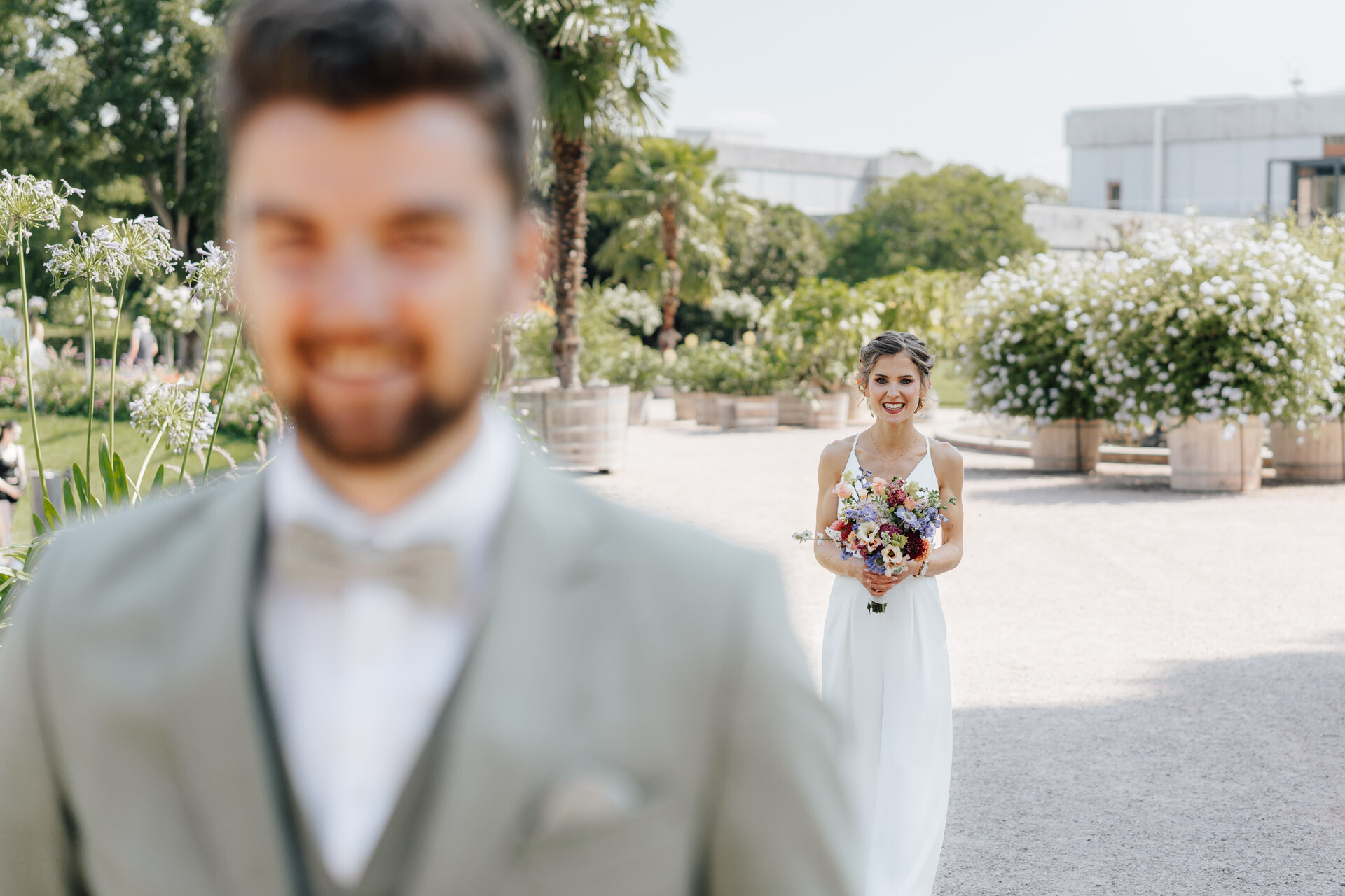 Eine Braut in einem weißen Kleid mit einem Blumenstrauß in der Hand lächelt, während sie an einem sonnigen Tag auf der Hochzeit Tante Polly Karlsruhe im Freien steht; üppiges Grün und weiße Blumen säumen den Weg, mit dem unscharfen Bräutigam in einem hellen Anzug im Vordergrund.