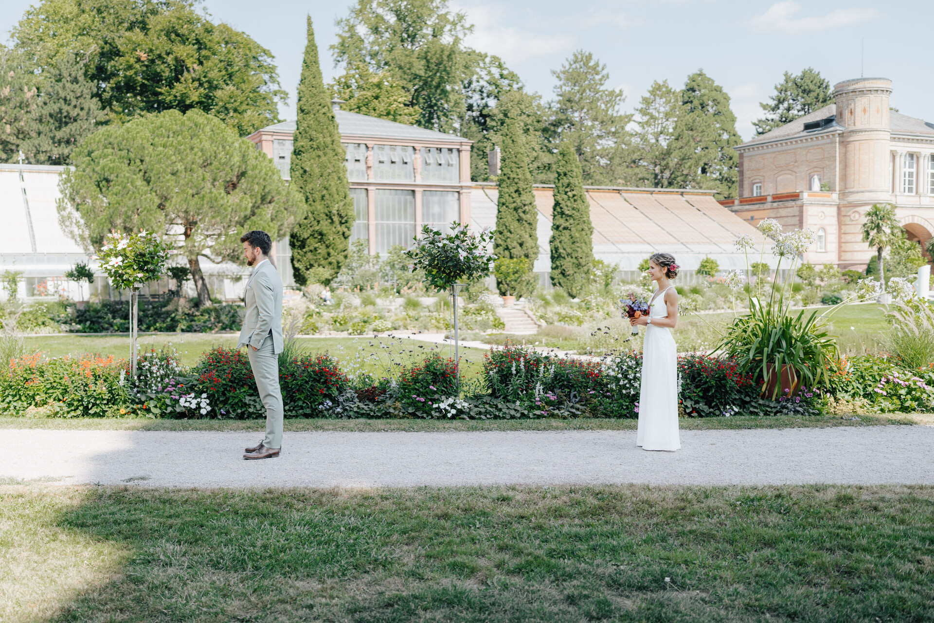 Eine Braut und ein Bräutigam stehen getrennt voneinander auf einem Gartenweg bei Hochzeit Tante Polly Karlsruhe und schauen sich an. Die Braut hält einen Blumenstrauß in der Hand, während im Hintergrund elegante Gewächshäuser und üppiges Grün zu sehen sind.