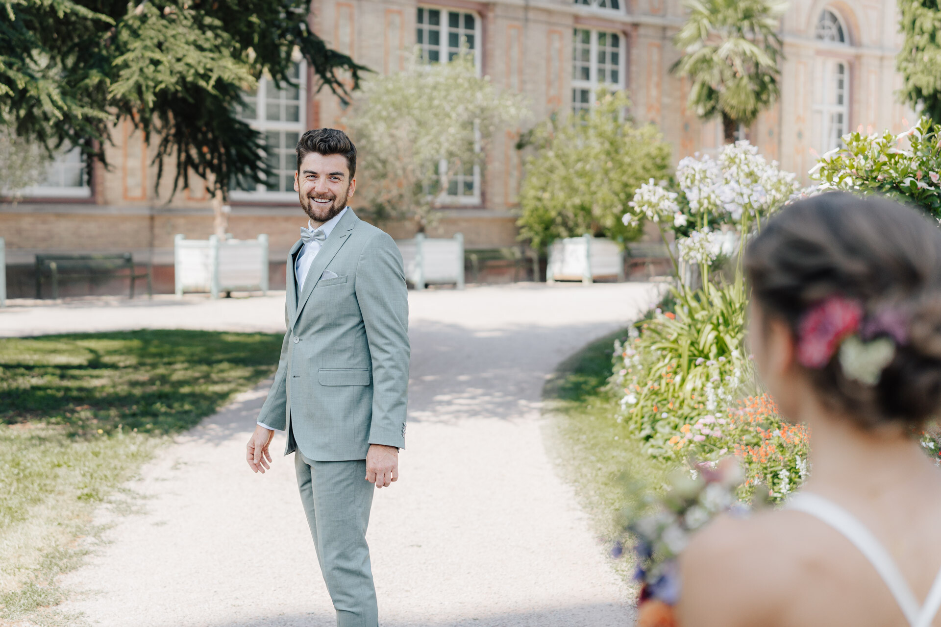 Ein Mann in einem hellblauen Anzug lächelt und blickt zurück auf eine Frau in einem weißen Kleid, die einen Blumenstrauß während ihrer Hochzeit bei Tante Polly Karlsruhe hält, umgeben von Gartenblumen und einem großen Gebäude im Hintergrund.