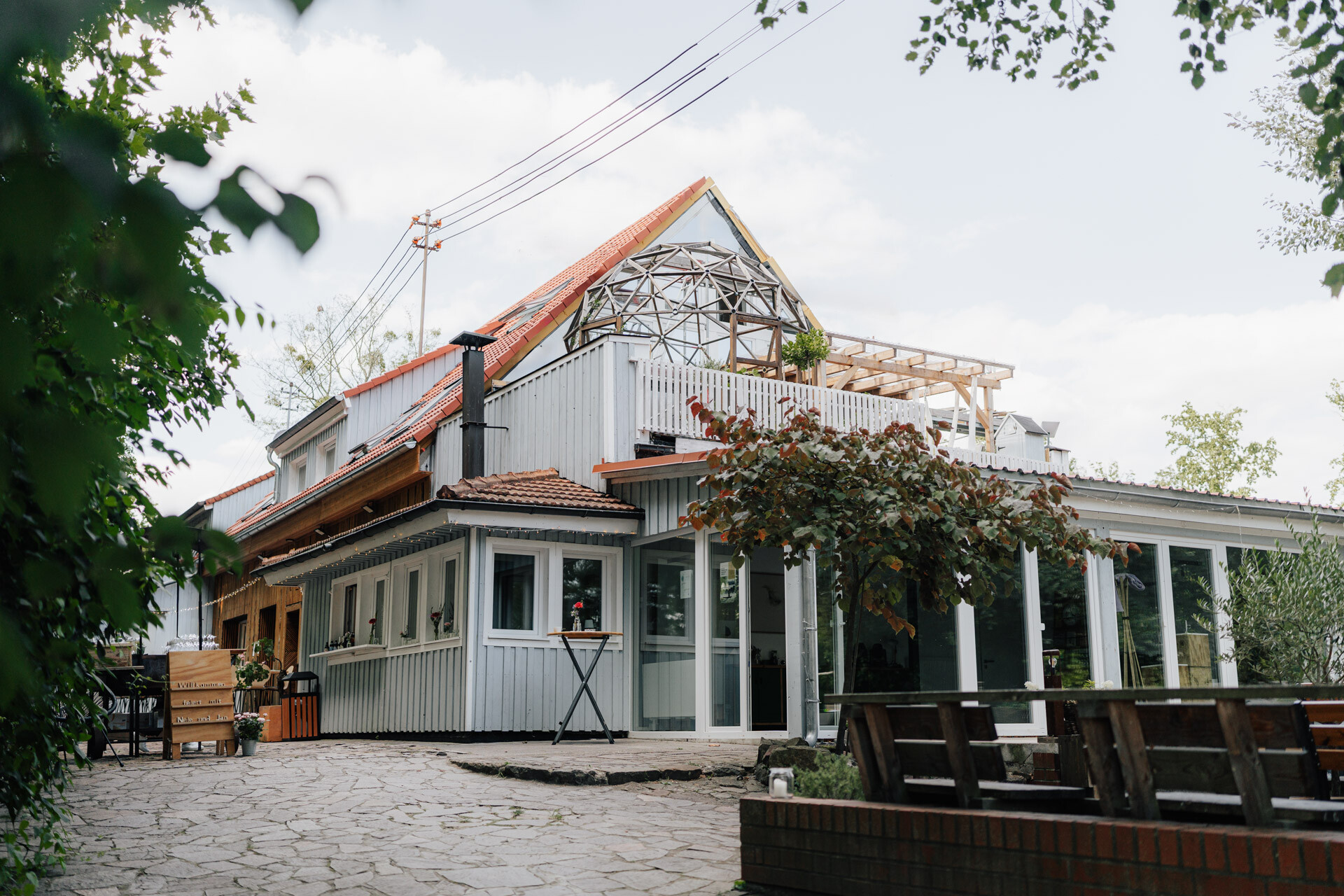 Ein modernes Haus mit großen Fenstern, einer Terrasse und einer Glaskuppel auf dem Dach, umgeben von Grün und Sitzgelegenheiten im Freien auf einer Steinterrasse - ideal für eine Hochzeit oder ein Treffen nach dem Vorbild von Tante Polly Karlsruhe.