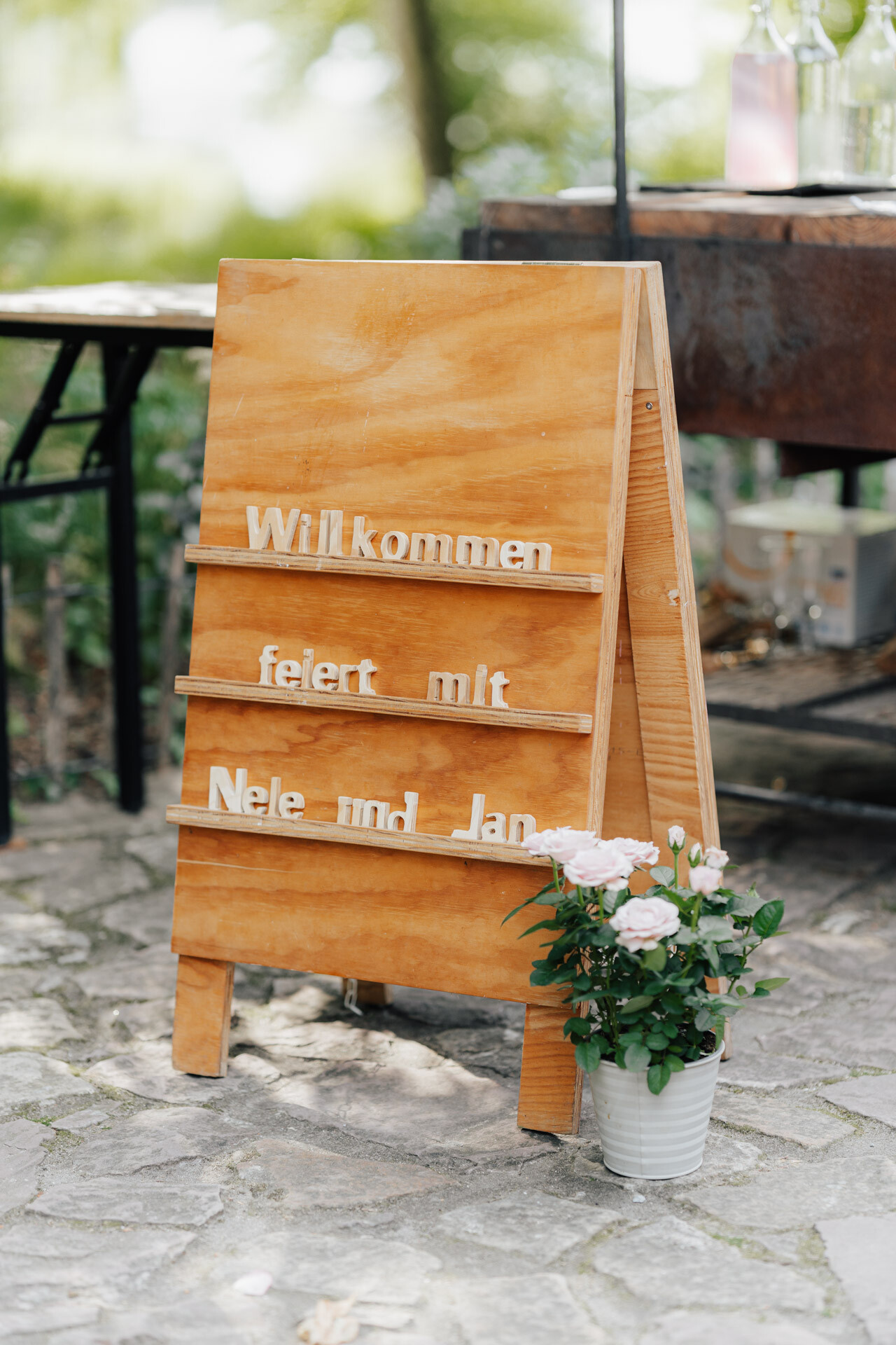 Auf einem hölzernen Schild steht "Willkommen feiert mit Nele und Jan" in großen Buchstaben. Es steht auf einem Steinweg neben einem weißen Topf mit rosafarbenen und weißen Rosen, inmitten von Tischen im Freien und Grünpflanzen bei Hochzeit Tante Polly Karlsruhe.