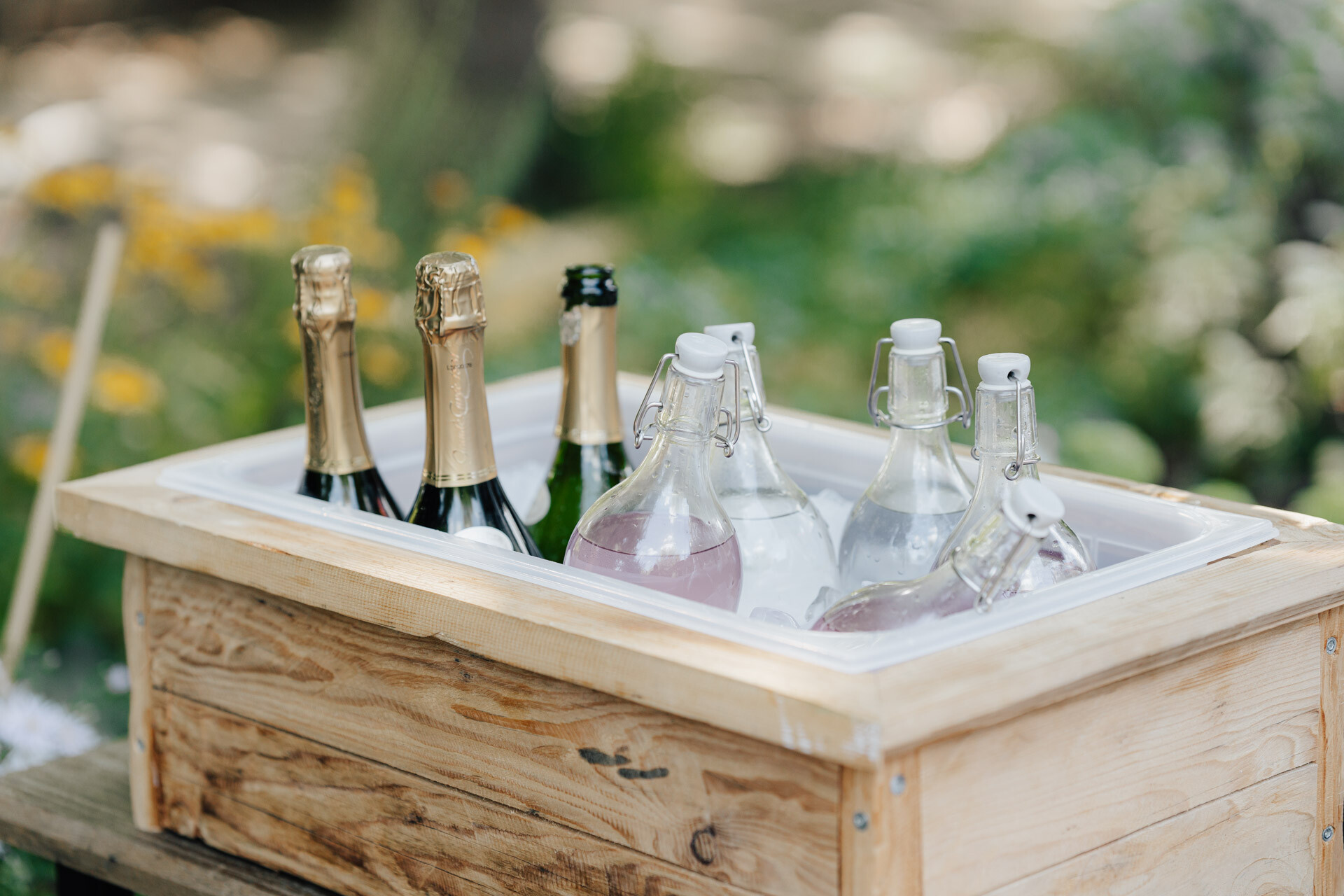 Eine Holzkiste, gefüllt mit Sektflaschen und gläsernen Bügelflaschen mit klaren und hellrosa Getränken, perfekt für eine Hochzeit bei Tante Polly in Karlsruhe, steht im Freien mit verschwommenen Grünpflanzen und Blumen im Hintergrund.