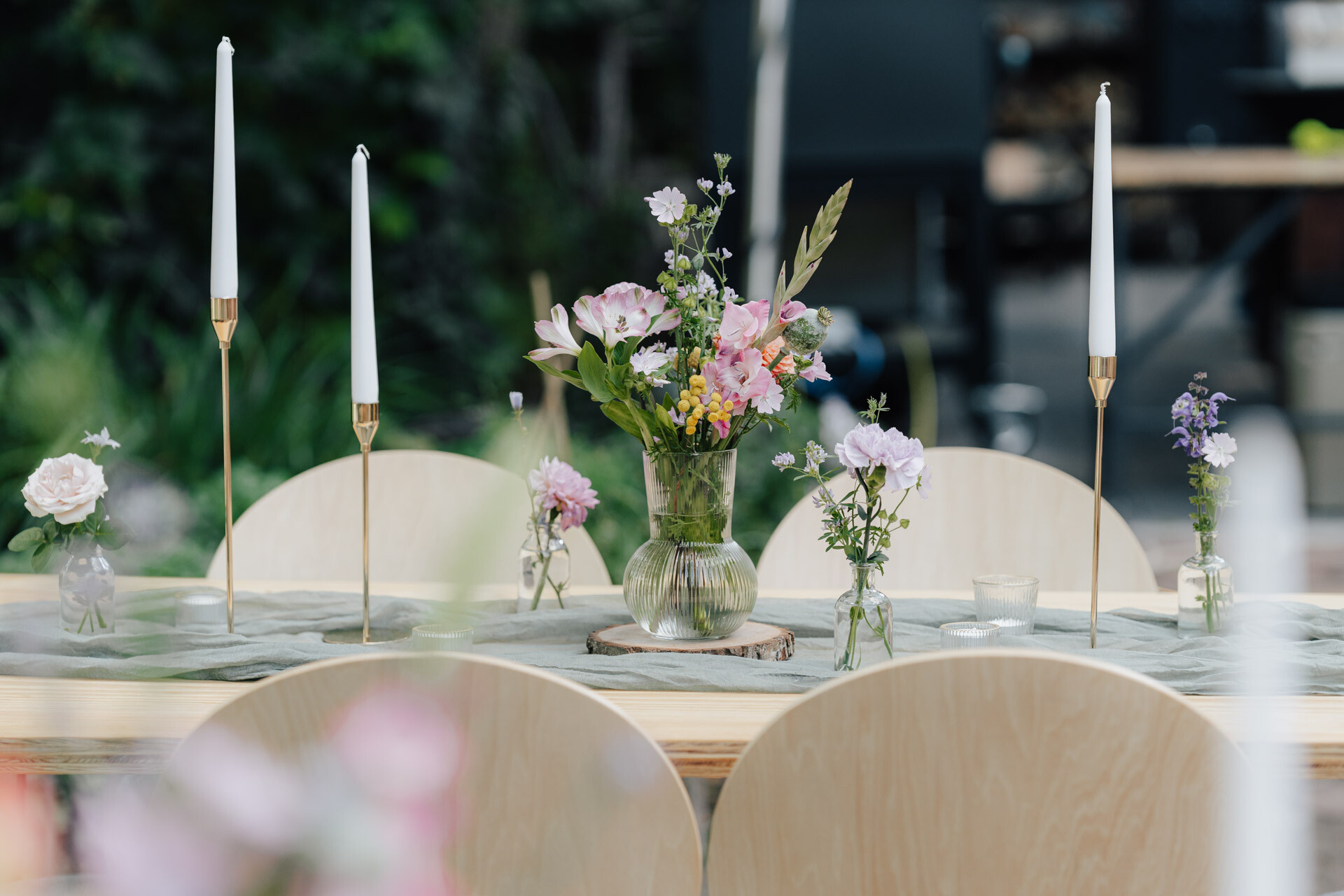 Ein Holztisch im Freien bei Hochzeit Tante Polly Karlsruhe, geschmückt mit einer Glasvase mit pastellfarbenen Blumen, umgeben von vier hellen Holzstühlen und hohen weißen Kerzen in goldenen Haltern. Unscharfes Grün vervollständigt die charmante Szene.