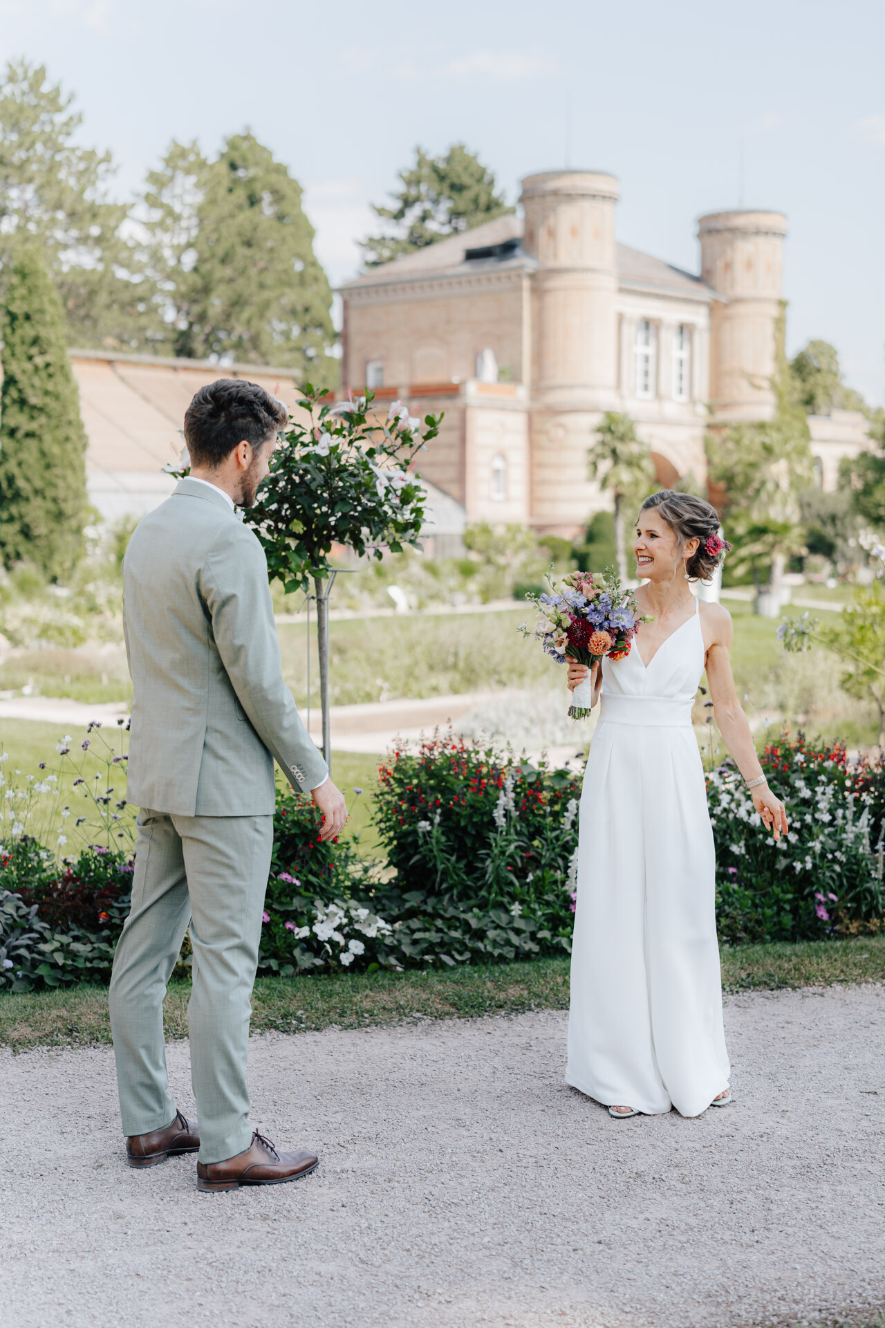 Eine Braut in einem weißen Kleid mit einem Blumenstrauß in der Hand lächelt einen Bräutigam in einem hellen Anzug an. Sie stehen im Freien in einem Garten bei Hochzeit Tante Polly Karlsruhe, mit Blumen, Grünzeug und einem großen historischen Gebäude im Hintergrund.