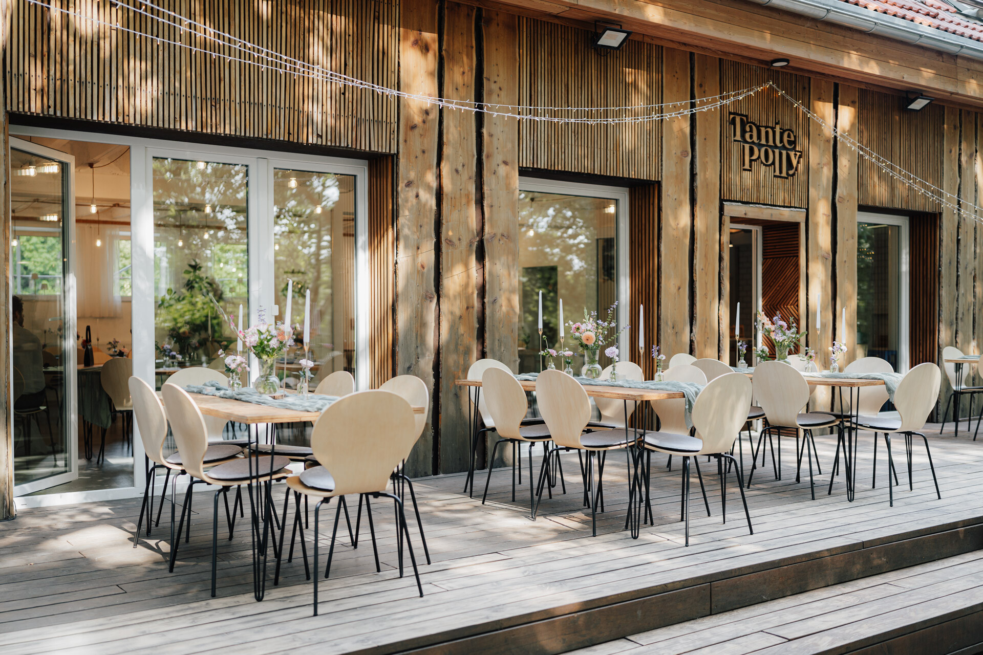 Ein Essbereich im Freien mit Holztischen und hellen Stühlen befindet sich auf einer Holzterrasse vor einem rustikalen Gebäude. Perfekt für eine Hochzeit, empfängt Tante Polly Karlsruhe seine Gäste mit Blumen, Kerzen und einem charmanten Schild über dem Eingang.