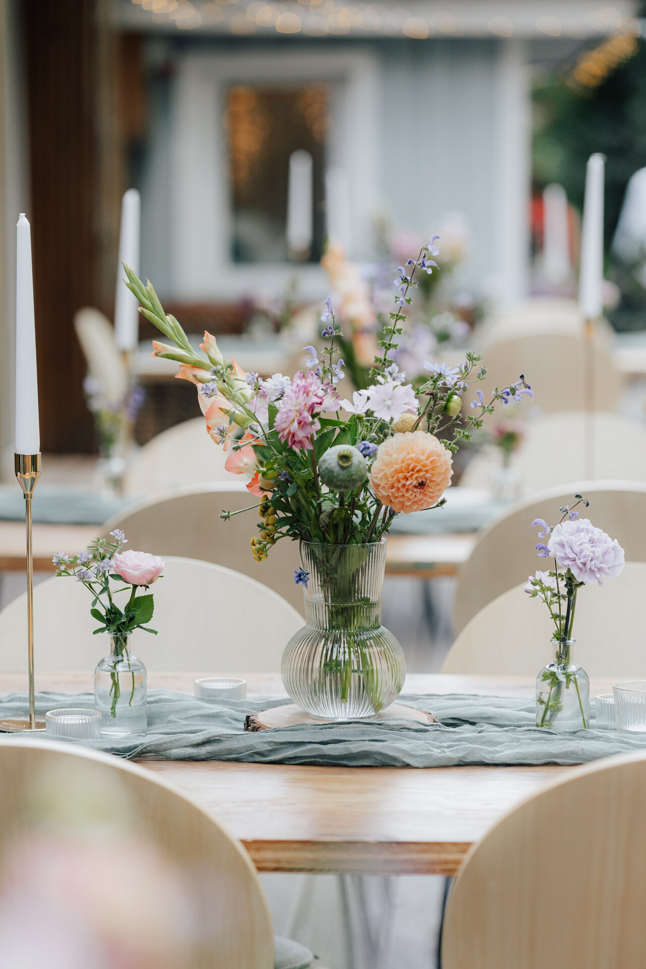 Ein Holztisch bei Hochzeit Tante Polly Karlsruhe ist mit einer Glasvase mit farbenfrohen Blumen, kleinen Gläsern mit einzelnen Blüten und hohen weißen Kerzen dekoriert und bietet einen eleganten und luftigen Rahmen für eine Veranstaltung.