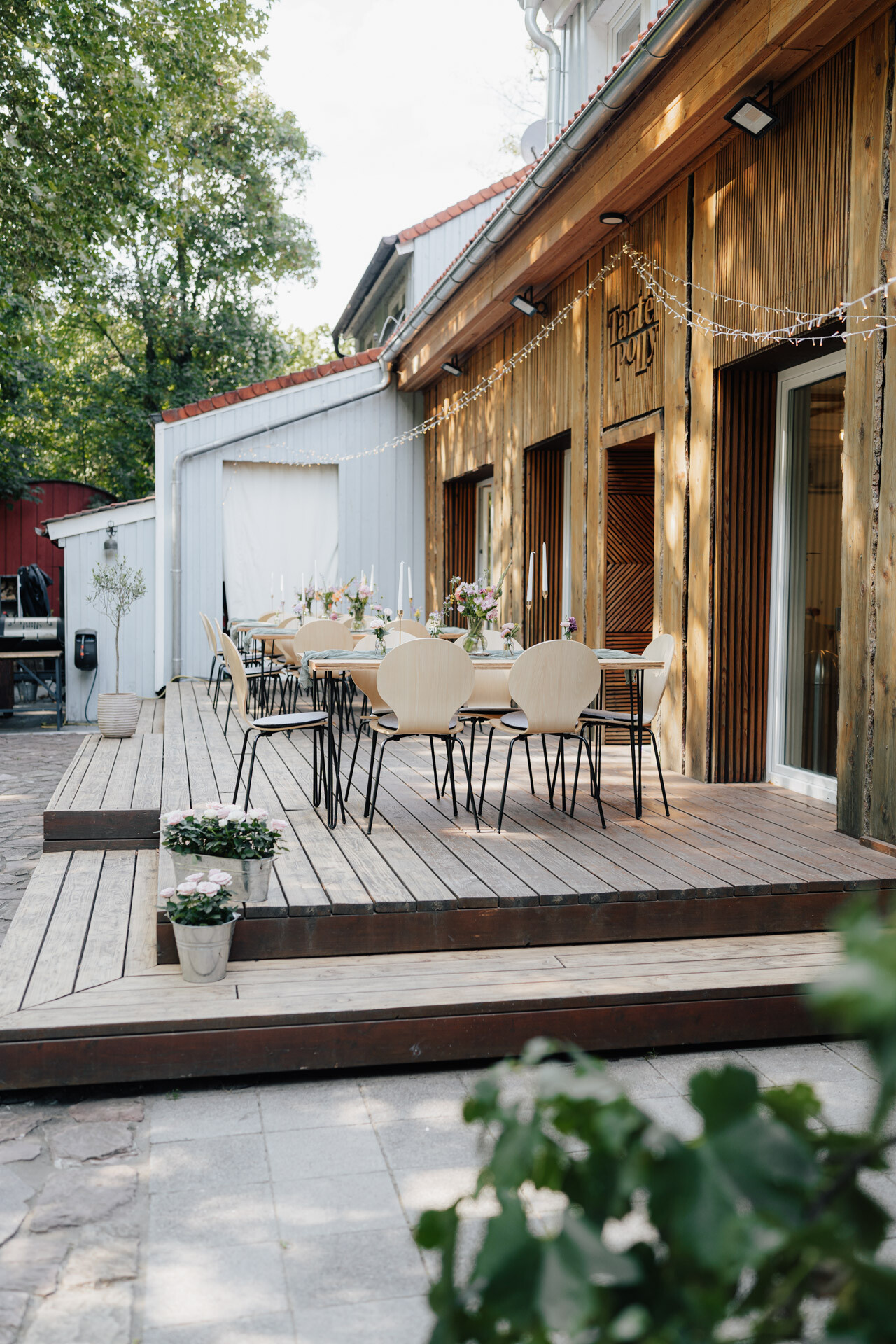 Eine hölzerne Außenterrasse mit modernen Stühlen und Tischen zum Essen, dekoriert mit Blumen und Lichterketten - perfekt für eine Hochzeit bei Tante Polly Karlsruhe, die sich neben einem rustikalen Gebäude inmitten von üppigem Grün befindet.