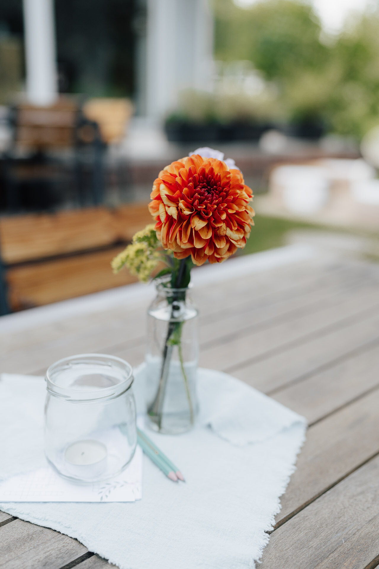 Eine einzelne orangefarbene Dahlienblüte in einer Glasvase steht auf einem Holztisch im Freien bei Hochzeit Tante Polly Karlsruhe, neben einer Kerze in einem Glas, zwei Buntstiften und einer weißen Stoffserviette. Der Hintergrund besteht aus unscharfem Grün und Terrassenmöbeln.