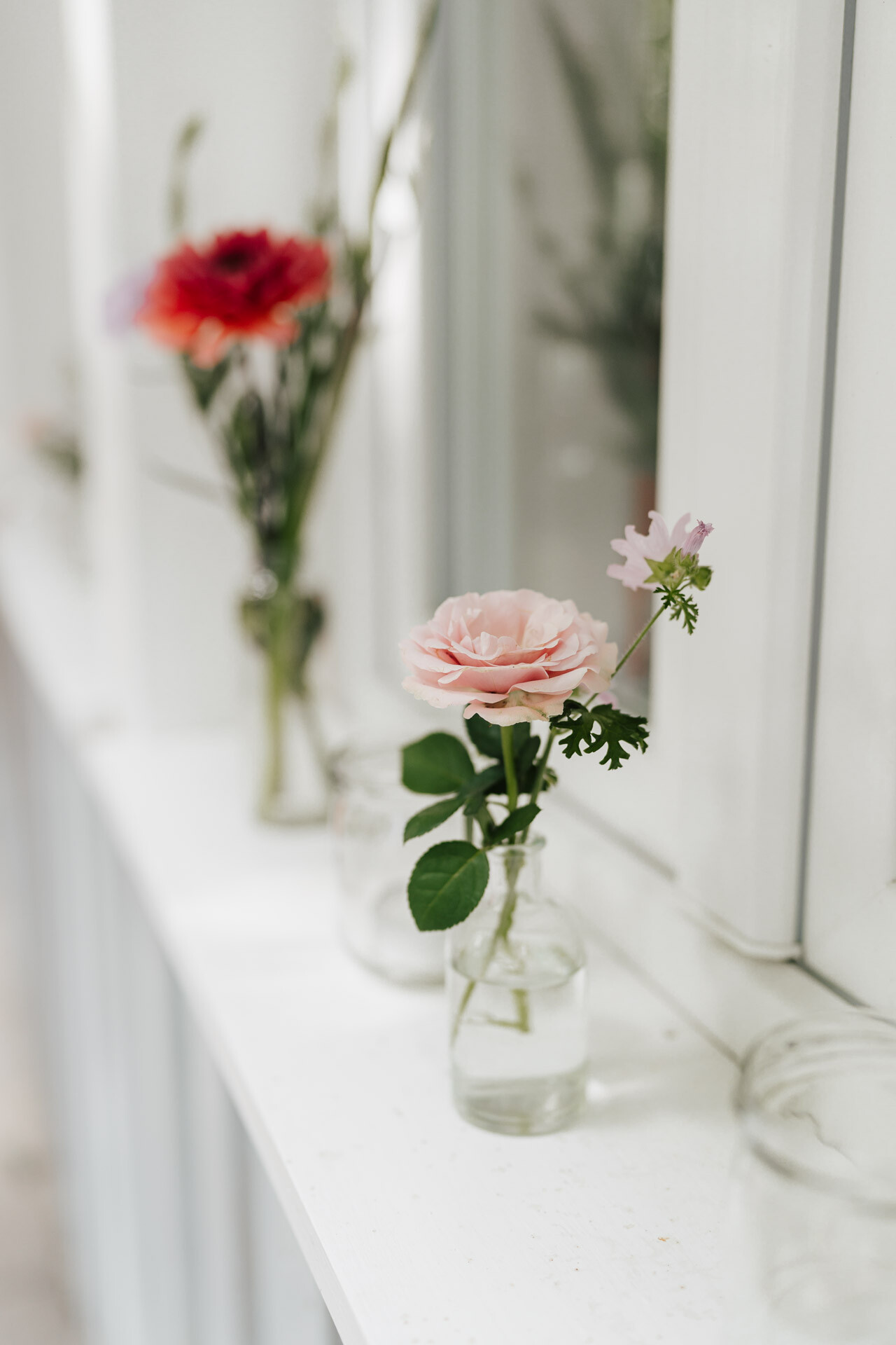 Ein kleines Glasgefäß mit einer hellrosa Rose und Grünzeug steht auf einer weißen Fensterbank bei Hochzeit Tante Polly Karlsruhe. Im Hintergrund ist ein weiteres Glas mit einer roten Blume leicht unscharf, was die helle und luftige Szene noch verstärkt.