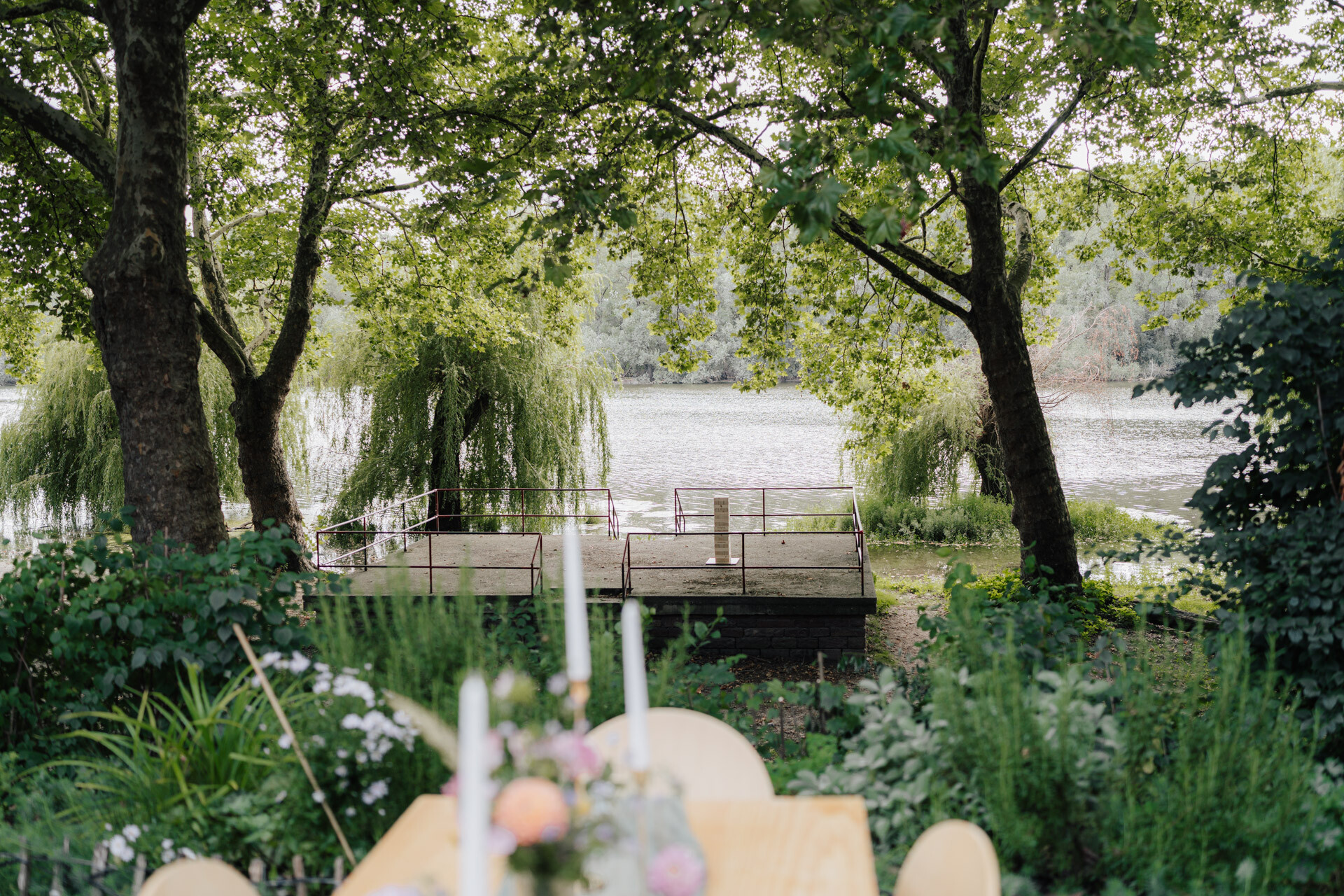 Ein hölzerner Steg erstreckt sich über einen ruhigen See, der von üppig grünen Bäumen umgeben ist. Im Vordergrund ist ein mit Blumen und Kerzen gedeckter Tisch, perfekt für eine Hochzeit bei Tante Polly Karlsruhe, zwischen Gartenpflanzen teilweise sichtbar.