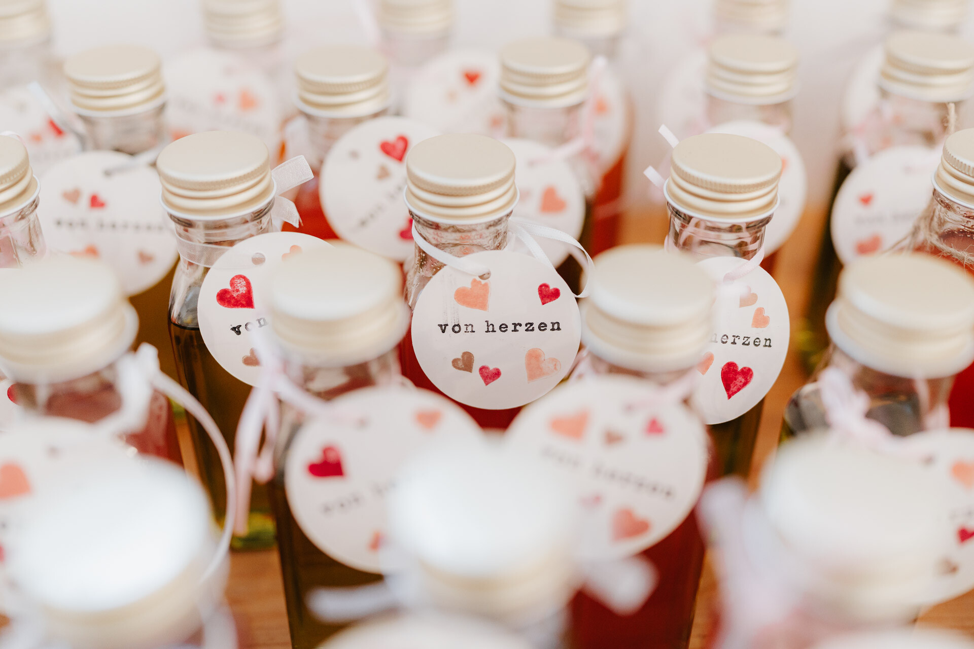 Mehrere Glasflaschen mit goldenen Verschlüssen, die jeweils mit einem runden Anhänger mit Herzmotiven und dem Schriftzug von herzen verziert sind, stehen bei Tante Polly Karlsruhe dicht beieinander - perfekt für eine Hochzeit.