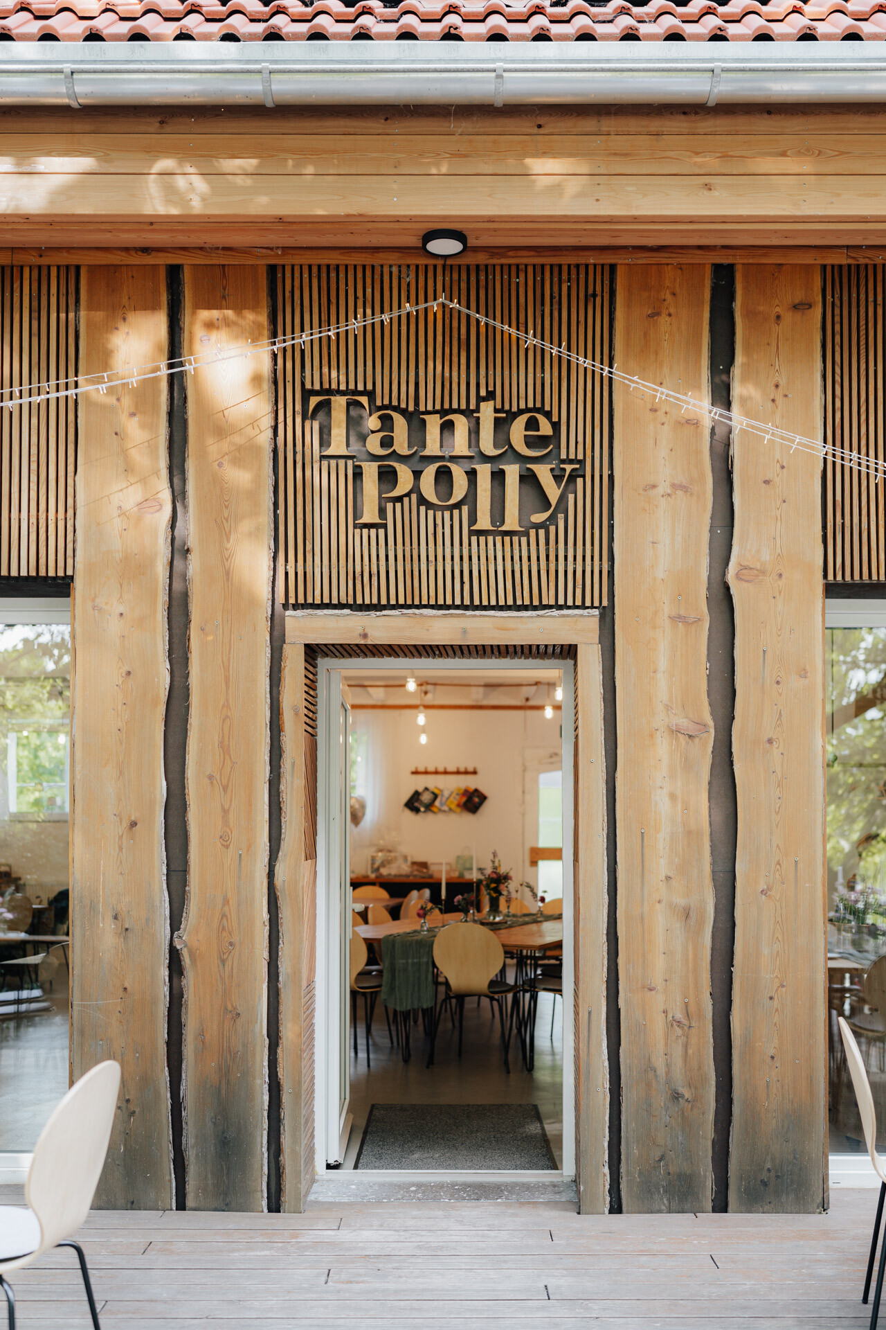 Eingang zur Hochzeit Tante Polly Karlsruhe - ein gemütliches Café mit hölzernen Außenwänden und einer offenen Tür, die zu einem warm beleuchteten Innenraum mit einladenden Esstischen und Stühlen führt.