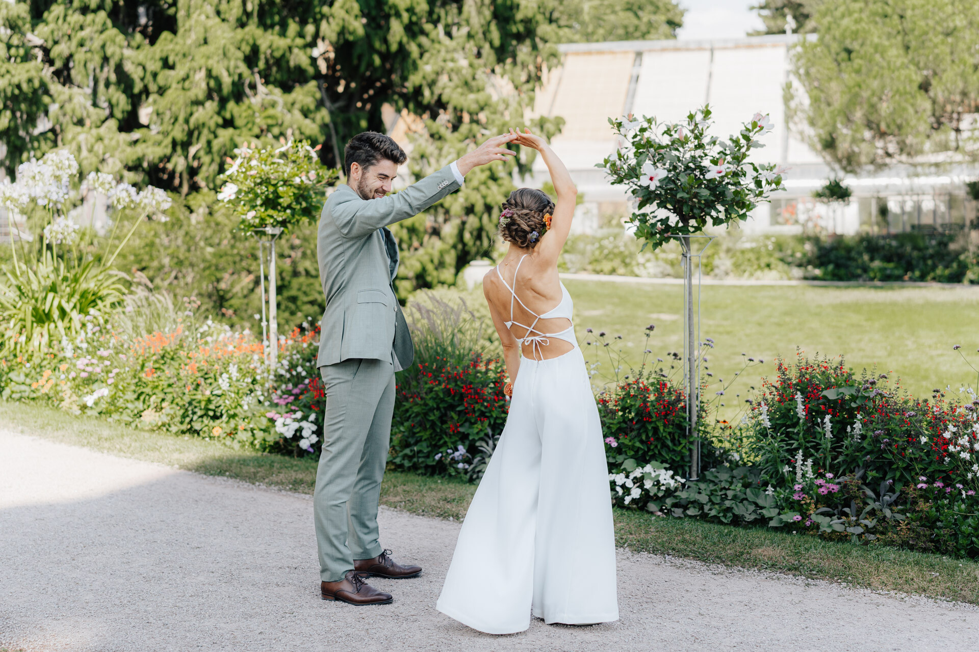 Ein Mann in einem hellgrünen Anzug wirbelt eine Frau in einem weißen, rückenfreien Kleid im Freien auf einem Gartenweg bei Hochzeit Tante Polly Karlsruhe, umgeben von Blumen und Grün an einem sonnigen Tag.