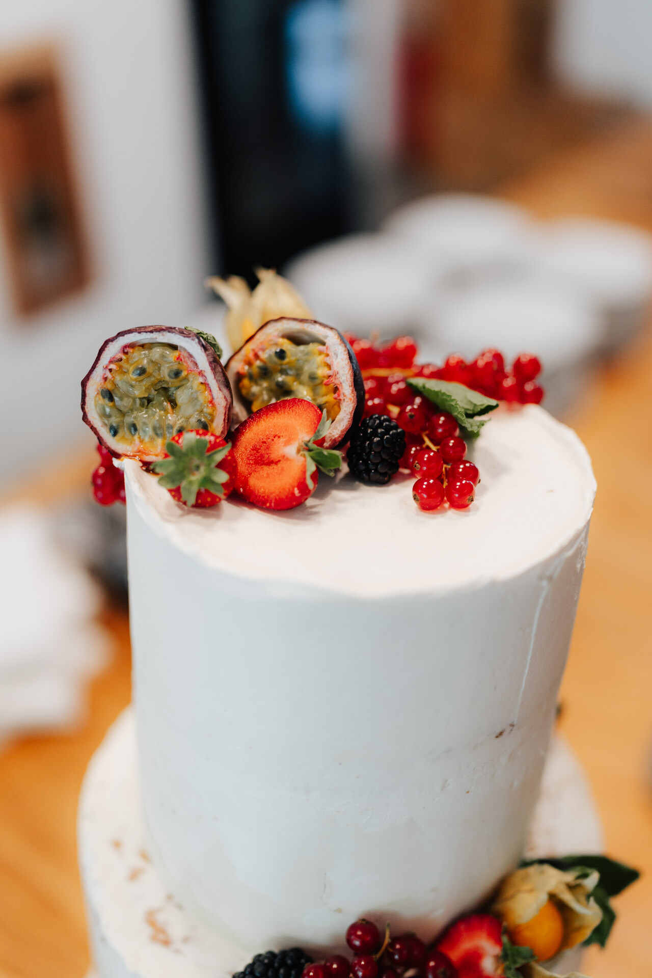 Eine weiße, glasierte, zweistöckige Torte von Hochzeit Tante Polly Karlsruhe, verziert mit halbierten Passionsfrüchten, Erdbeeren, roten Johannisbeeren, Brombeeren und grünen Blättern, elegant auf einem Holztisch präsentiert.