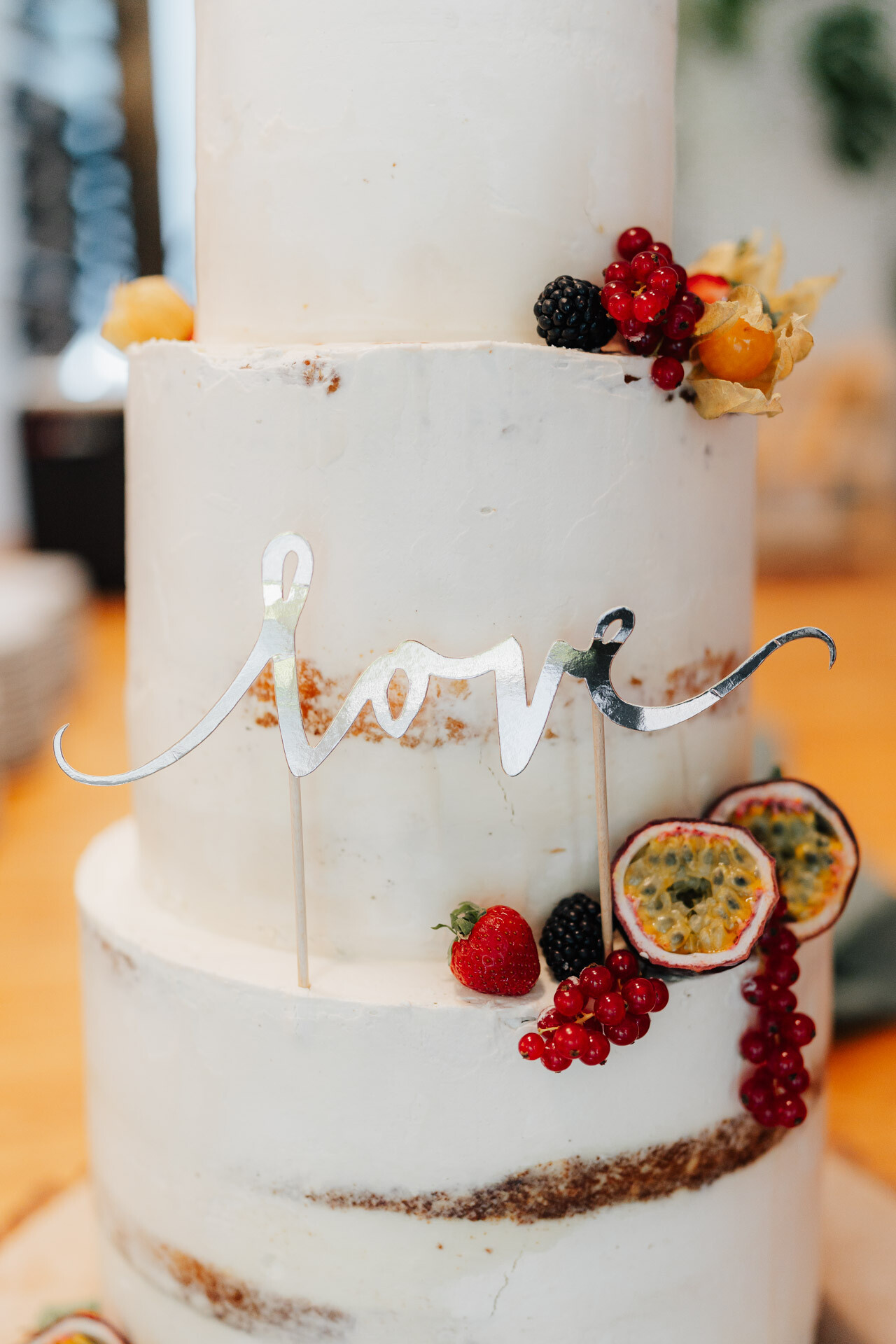 Eine dreistöckige weiße Torte, dekoriert mit verschiedenen frischen Beeren und Passionsfruchtscheiben, gekrönt von einem silbernen Schriftzug mit der Aufschrift Liebe - perfekt für eine Hochzeit bei Tante Polly Karlsruhe.