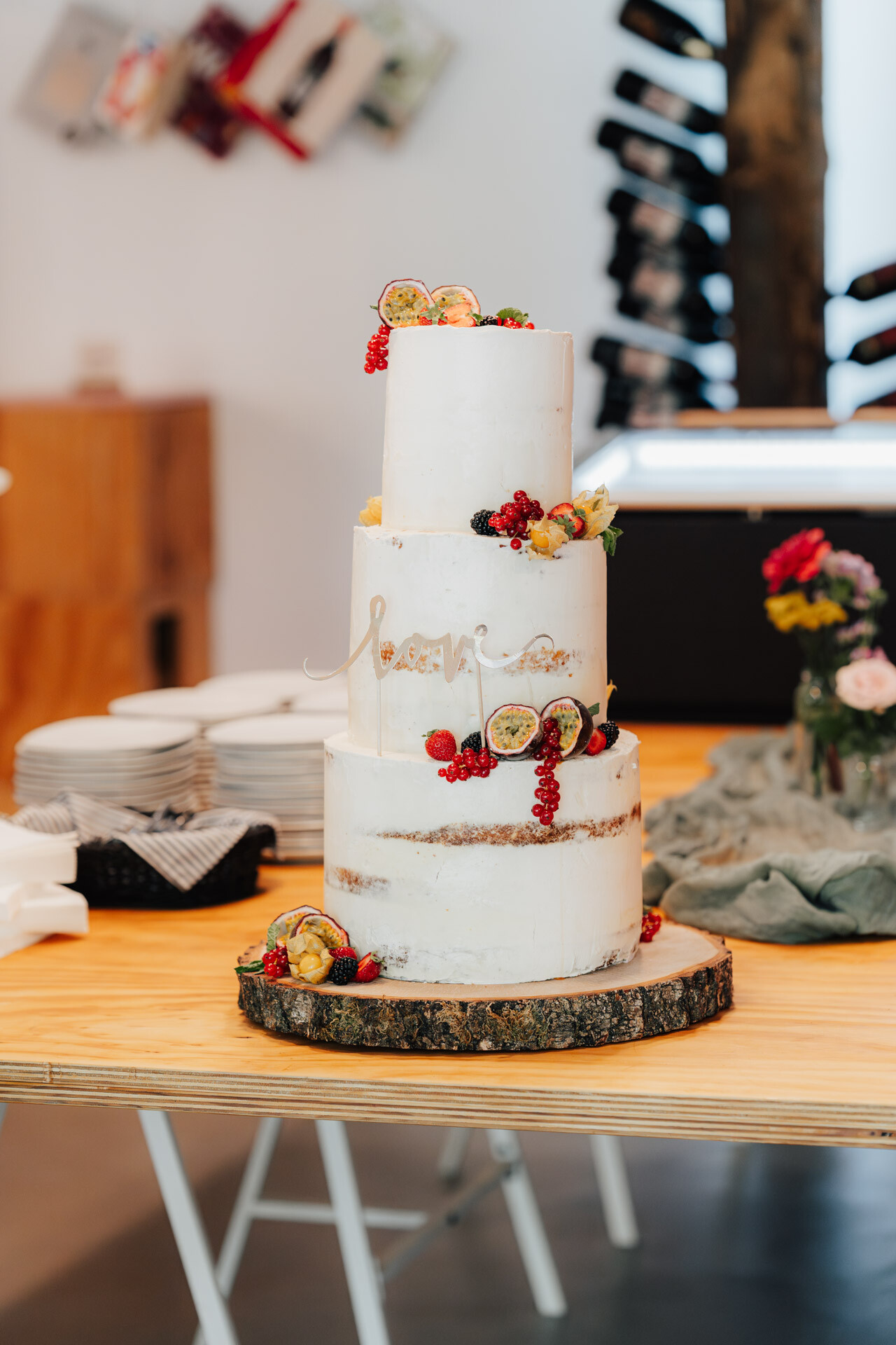 Eine dreistöckige weiße Hochzeitstorte, dekoriert mit frischen Beeren und geschnittenen Passionsfrüchten, steht bei Hochzeit Tante Polly Karlsruhe auf einer Holzplatte, umgeben von Tellern, Besteck, Blumen und Weinflaschen im Hintergrund.