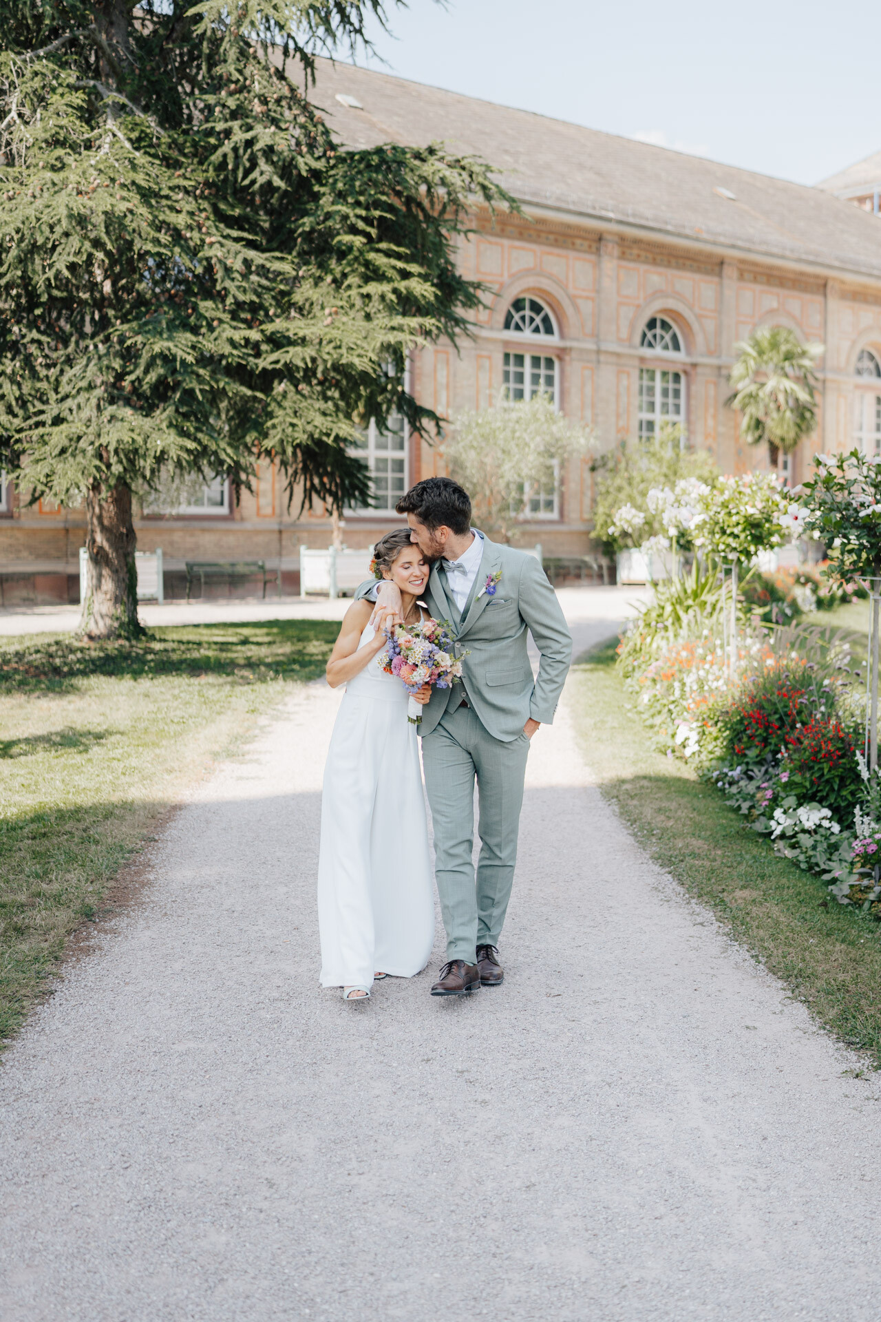 Eine Braut in einem weißen Kleid und ein Bräutigam in einem hellgrauen Anzug gehen Arm in Arm einen Gartenweg bei Hochzeit Tante Polly Karlsruhe entlang, lächelnd und liebevoll, mit Blumen und einem historischen Gebäude im Hintergrund.