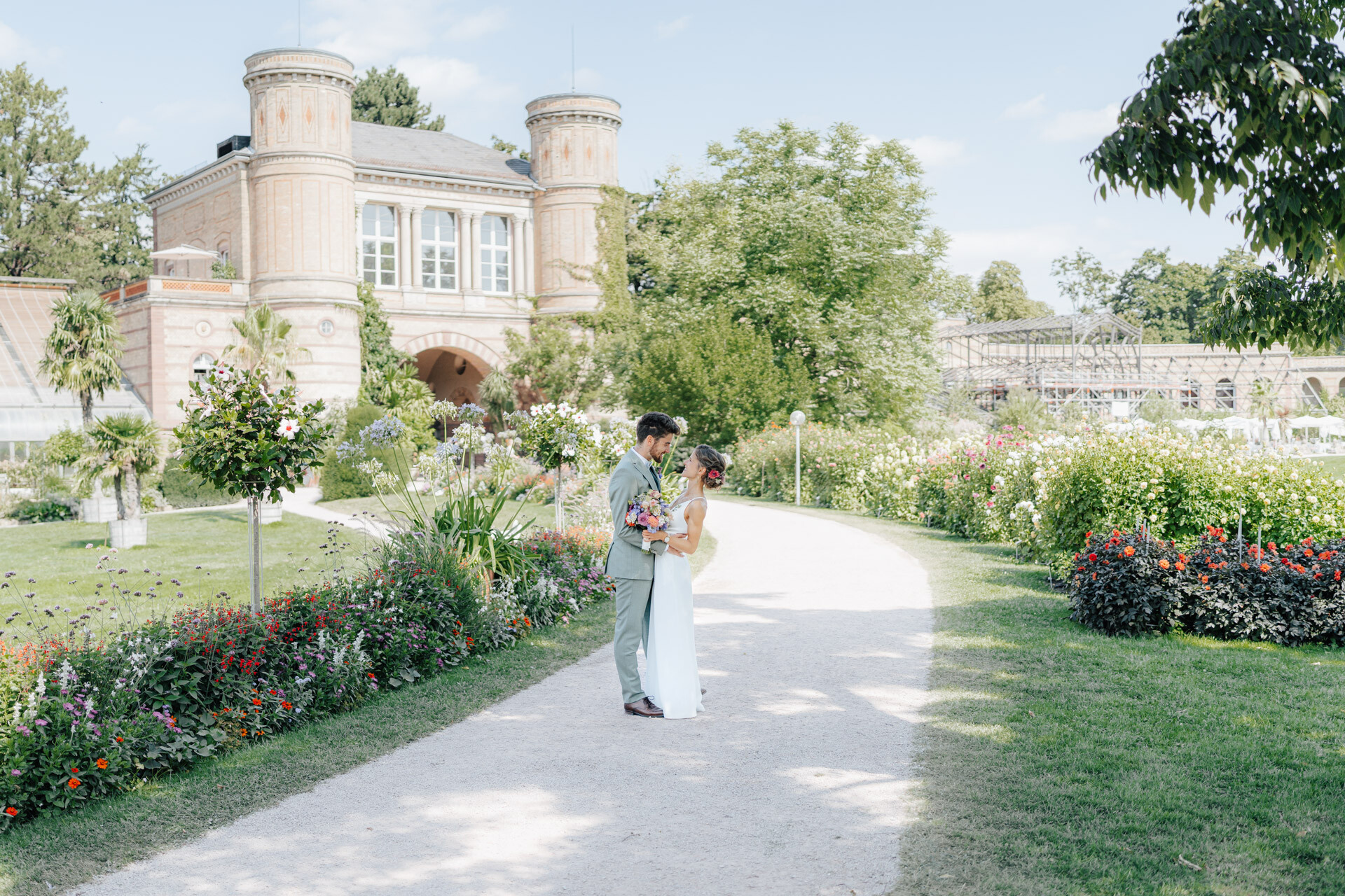 Braut und Bräutigam umarmen sich auf einem sonnenbeschienenen, von Blumen gesäumten Gartenweg bei Hochzeit Tante Polly Karlsruhe, mit einem historischen Backsteingebäude im Hintergrund unter einem klaren blauen Himmel.