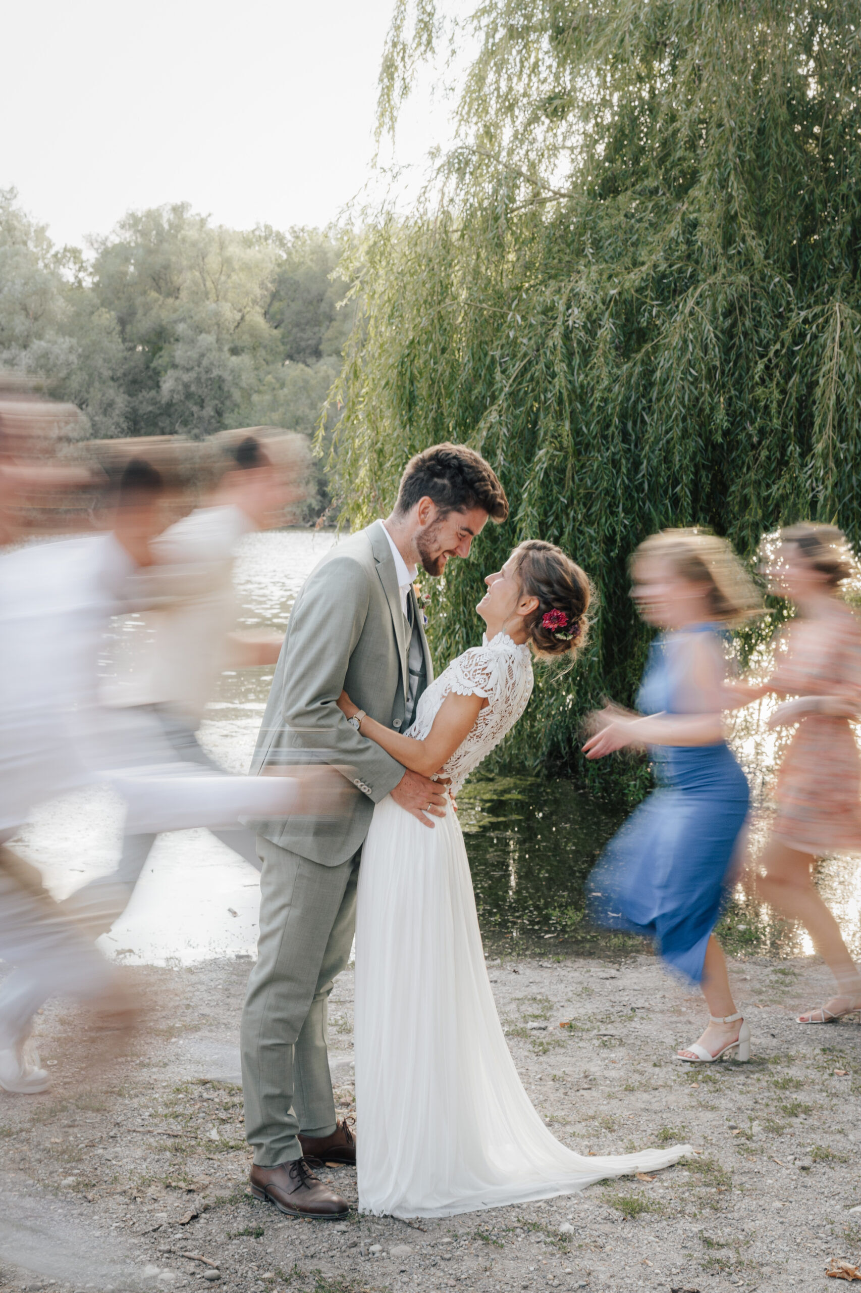 Braut und Bräutigam lächeln sich an und umarmen sich am Ufer eines Sees bei Hochzeit Tante Polly Karlsruhe, während Gäste in farbenfrohen Outfits an ihnen vorbeiziehen und eine lebhafte, fröhliche Szene schaffen. Im Hintergrund schimmern Bäume und Wasser.