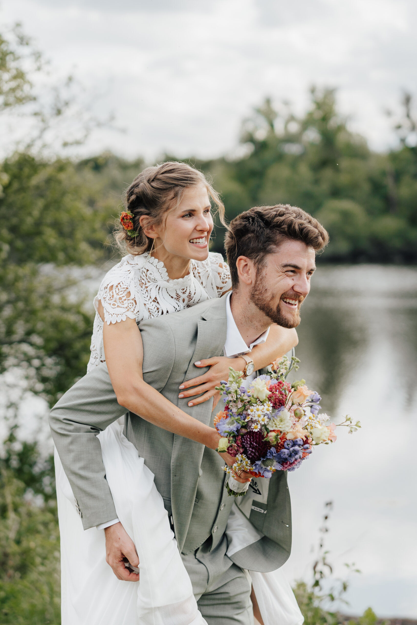 Eine lächelnde Braut in einem weißen Spitzenkleid reitet huckepack auf einem lachenden Bräutigam in einem hellgrauen Anzug, der einen bunten Blumenstrauß in der Hand hält, auf ihrer Hochzeit Tante Polly Karlsruhe, mit Grünpflanzen und einem ruhigen See im Hintergrund.
