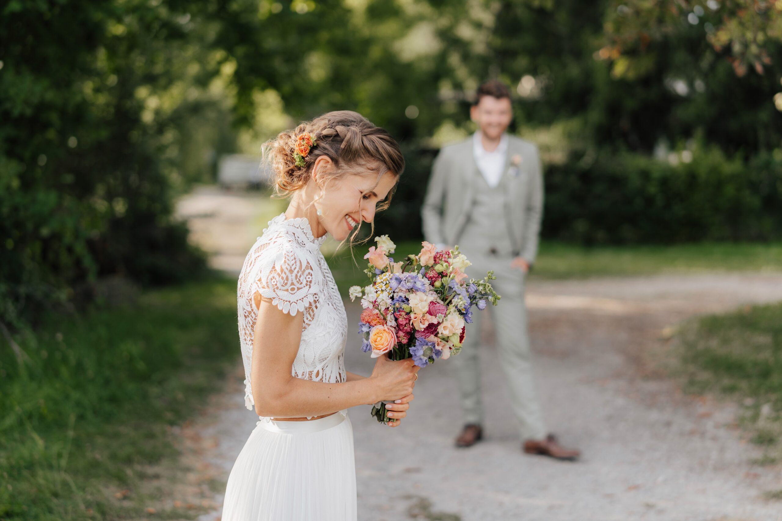 Eine Braut in einem weißen Kleid strahlt mit einem bunten Blumenstrauß auf einem von Bäumen gesäumten Weg bei Hochzeit Tante Polly Karlsruhe, während ein Mann in einem hellen Anzug, wahrscheinlich der Bräutigam, unscharf im Hintergrund steht und sie anlächelt.