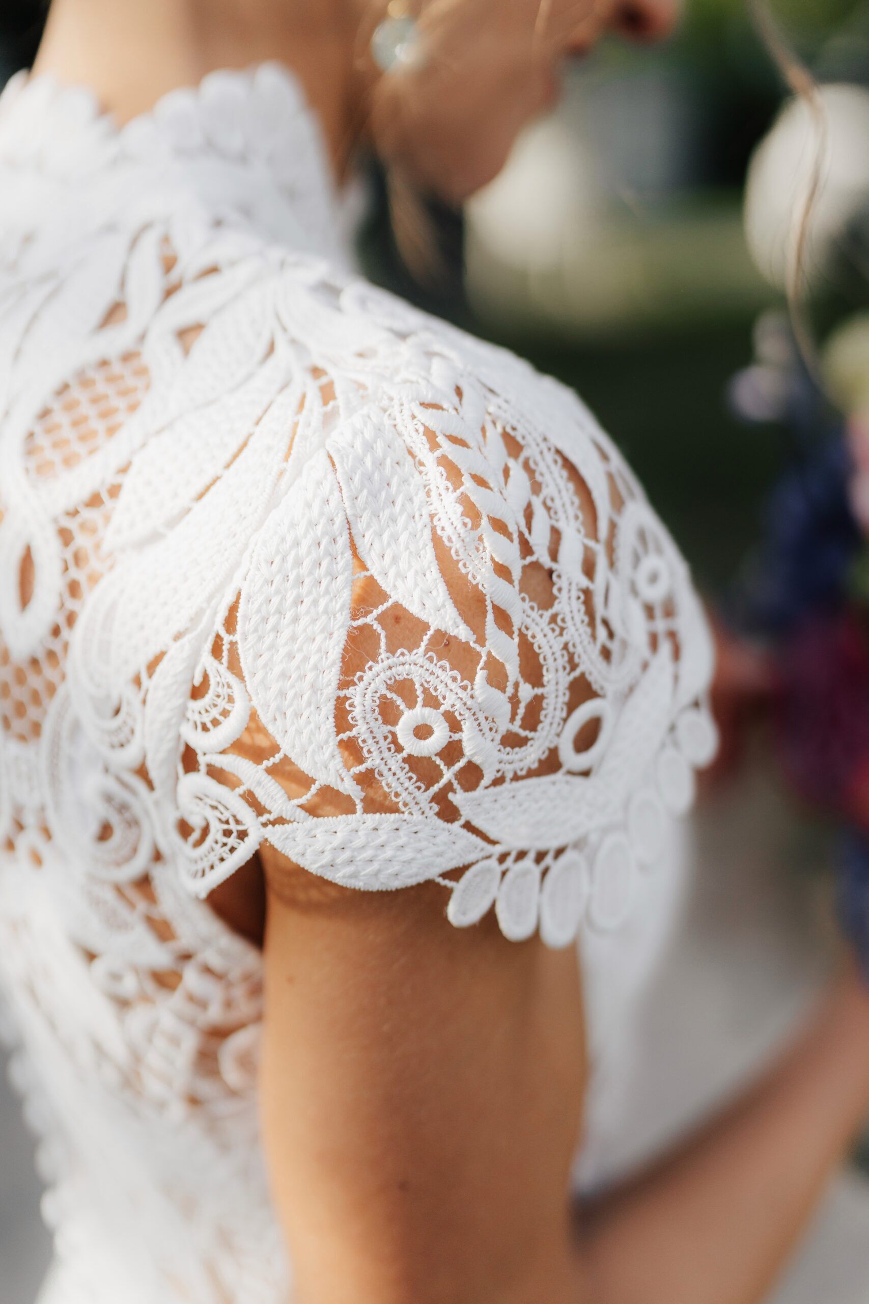 Nahaufnahme der Schulter einer Frau in einem weißen Spitzenkleid mit komplizierten Mustern und zarten Blumendetails, perfekt für eine Hochzeit bei Tante Polly Karlsruhe. Der unscharfe Hintergrund lenkt die Aufmerksamkeit auf die atemberaubende Textur und das Design der Spitze.