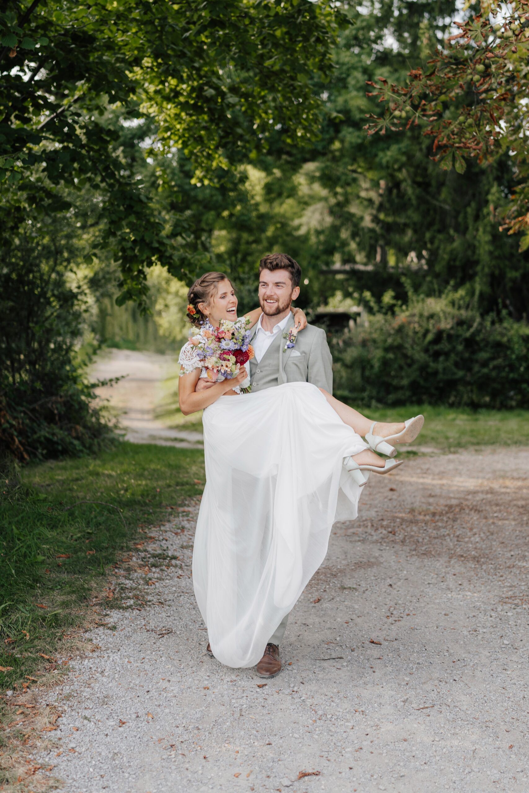Ein Bräutigam in einem hellen Anzug trägt eine lächelnde Braut in einem weißen Kleid, die einen Blumenstrauß in der Hand hält, auf einem von grünen Bäumen umgebenen Kiesweg bei Hochzeit Tante Polly Karlsruhe.