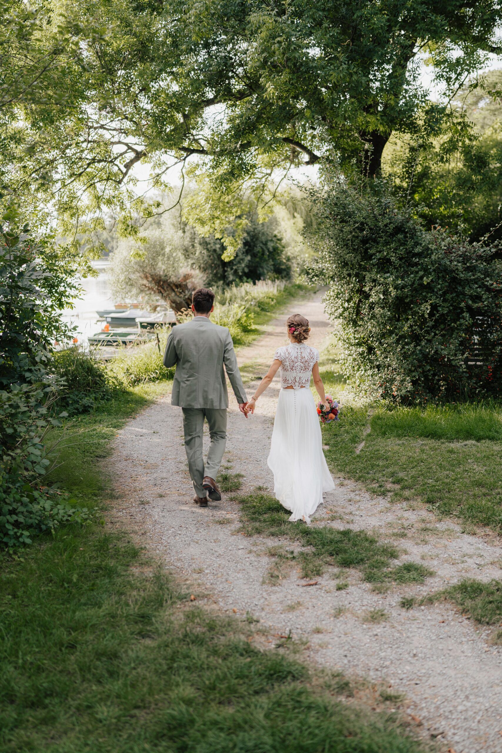 Ein Paar in Hochzeitskleidung schlendert Hand in Hand über einen von Bäumen gesäumten Kiesweg bei Hochzeit Tante Polly Karlsruhe. Die Braut in Weiß hält einen Blumenstrauß, und der Bräutigam im grauen Anzug ist an einem sonnigen Tag von üppigem Grün umgeben.