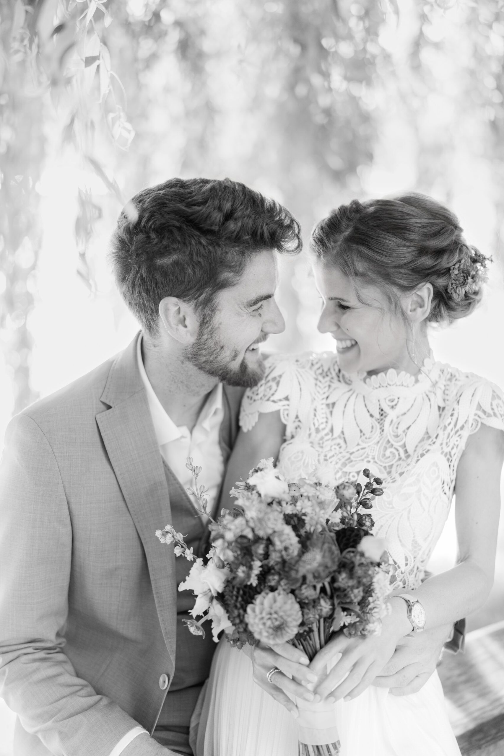 Eine Braut und ein Bräutigam sitzen eng beieinander und lächeln sich während ihrer Hochzeit bei Tante Polly Karlsruhe freudig an. Die Braut hält einen Blumenstrauß, beide tragen Hochzeitskleidung. Das schwarz-weiße Bild hat eine weiche, romantische Note.