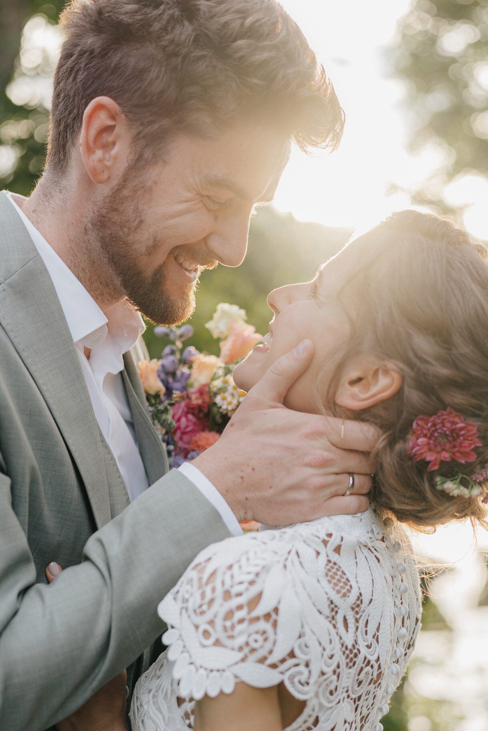 Ein glückliches Paar in Hochzeitskleidung lächelt und umarmt sich im Freien bei Hochzeit Tante Polly Karlsruhe, während die Sonne hinter ihnen scheint. Die Frau trägt ein Spitzenkleid und Blumen im Haar, während der Mann sanft ihr Gesicht hält, beide sehen fröhlich und verliebt aus.