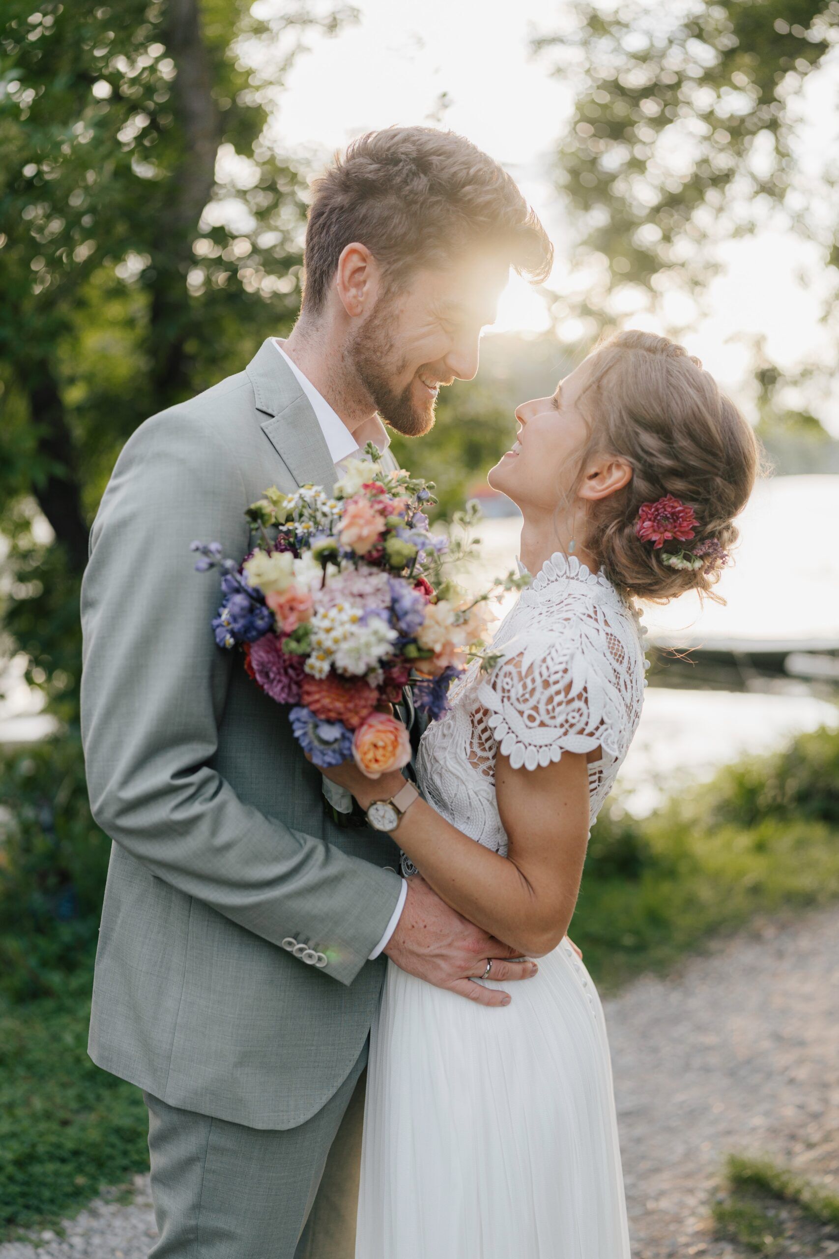 Eine Braut und ein Bräutigam umarmen sich im Freien bei Sonnenuntergang während ihrer Hochzeit Tante Polly Karlsruhe und lächeln sich an. Die Braut hält einen bunten Blumenstrauß in ihrem weißen Spitzenkleid, während der Bräutigam in einem hellgrauen Anzug unter sonnenbeschienenen Bäumen steht.
