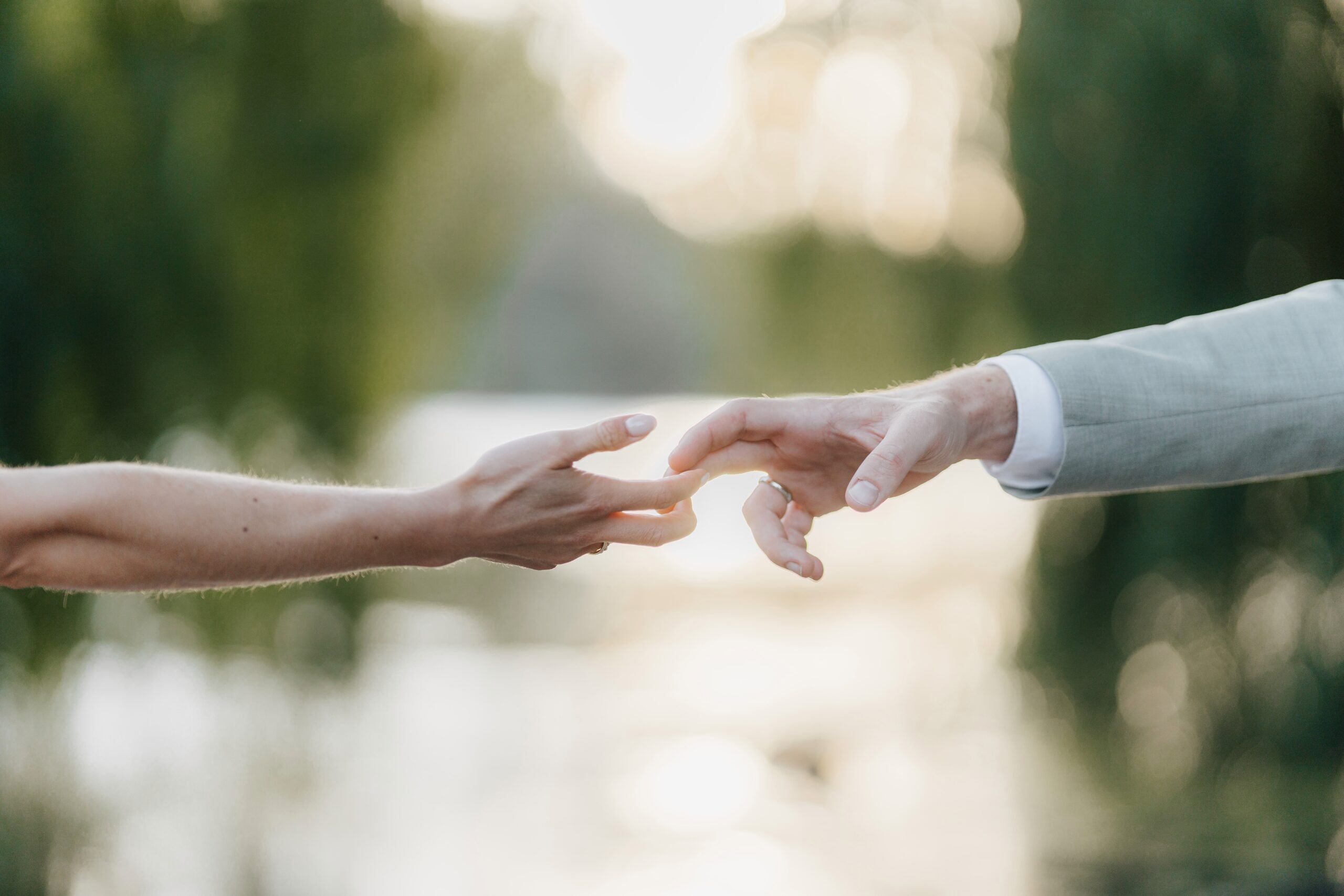 Zwei Hände, eine in einem leichten Anzugärmel und eine nackt, strecken sich einander entgegen, wobei sich die Finger sanft berühren - ein zärtlicher Moment auf einer Hochzeit bei Tante Polly Karlsruhe vor einem weichen, verschwommenen Hintergrund im Freien.