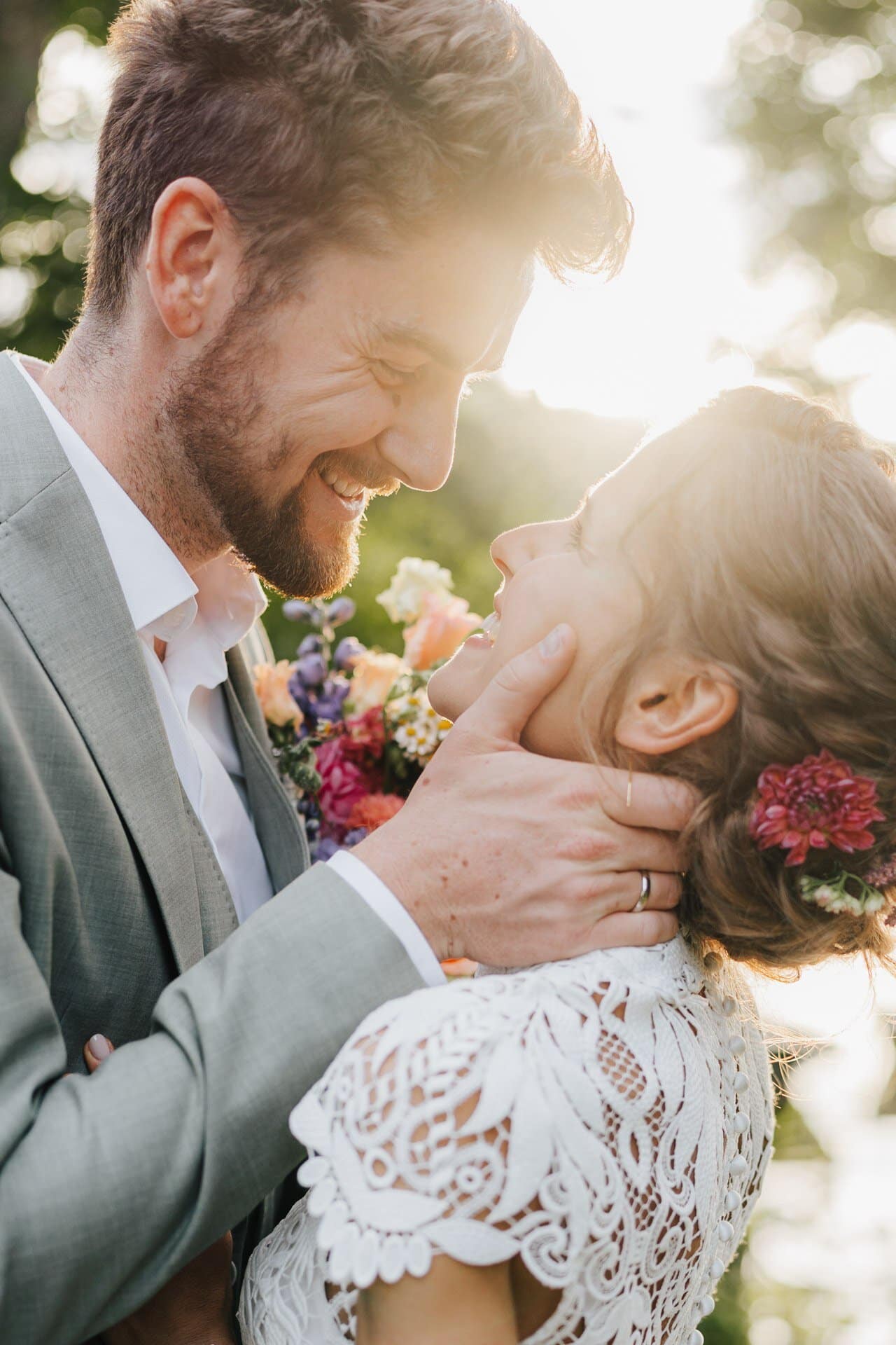 Ein Bräutigam in einem hellen Anzug lächelt und hält sanft das Gesicht einer Braut in einem Spitzenkleid mit Blumen im Haar. Aufgenommen von Hochzeitsfotograf Göppingen, blicken sie sich im Freien bei sanftem Sonnenlicht in die Augen.