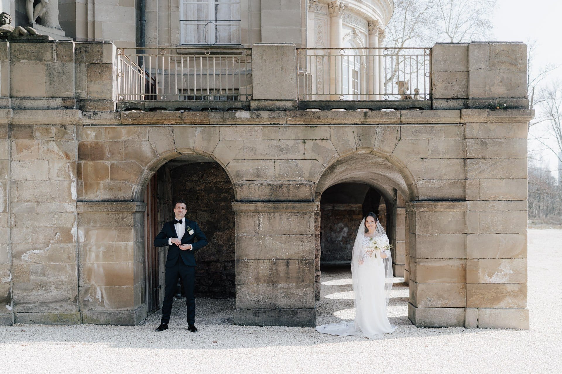 Eine Braut in weißem Kleid und Schleier steht unter einem steinernen Torbogen, während der Bräutigam in einem schwarzen Anzug unter einem anderen steht, wunderschön eingefangen von einem Hochzeitsfotografen Göppingen in einem historischen Gebäude mit gewölbten Eingängen.