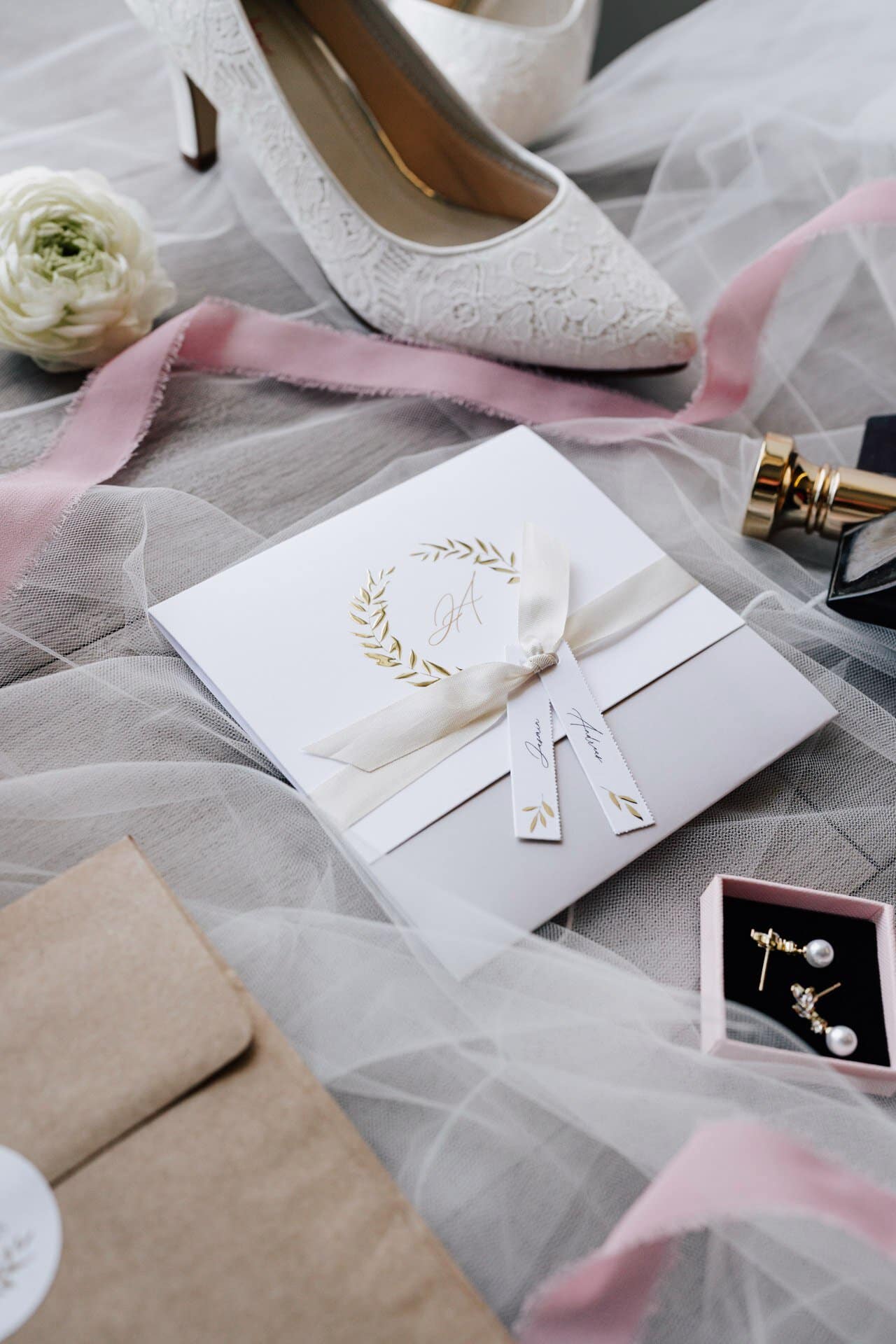 Eine weiße Hochzeitseinladung mit goldenem Monogramm und Schleife liegt auf durchsichtigem Stoff, umgeben von einem weißen Spitzenschuh, Parfüm, rosa Schleife, Ohrringen, einer Blume und einem braunen Umschlag - perfekt eingefangen von Hochzeitsfotograf Göppingen.