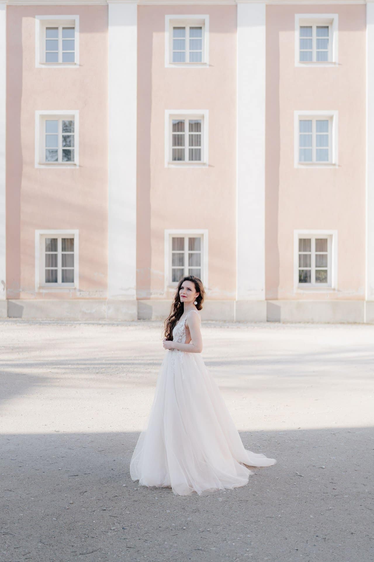 Eine Frau in einem fließenden weißen Kleid steht vor einem pastellrosa Gebäude mit weiß umrandeten Fenstern, das in sanftes Sonnenlicht getaucht ist. Die von Hochzeitsfotograf Göppingen aufgenommene Szene wirkt elegant und heiter.