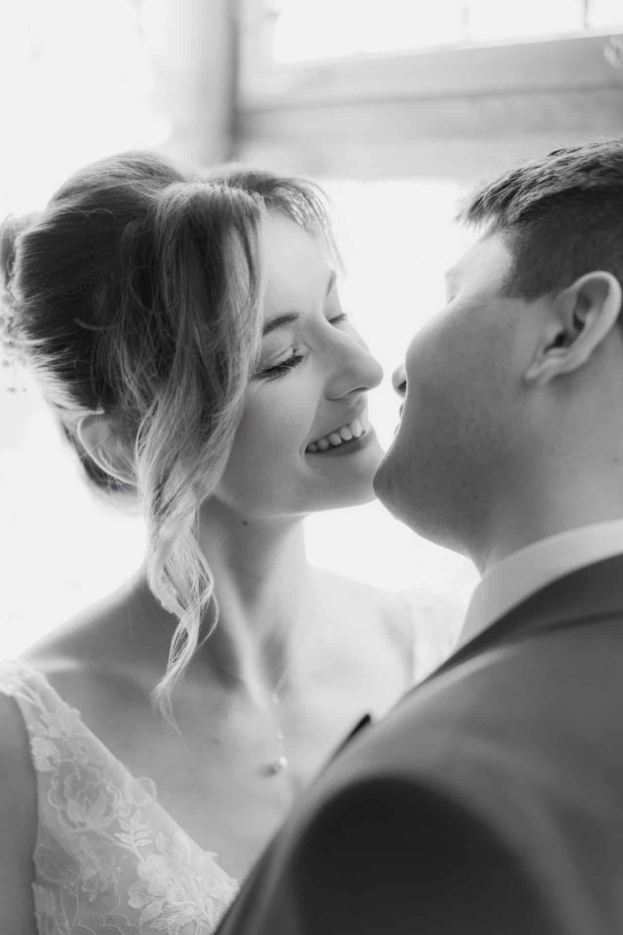 Ein Schwarz-Weiß-Foto von Hochzeitsfotograf Göppingen zeigt ein lächelndes Brautpaar, das sich eng umschlungen gegenübersteht und im Begriff ist, sich zu küssen. Die Braut trägt ein Spitzenkleid und der Bräutigam einen Anzug, beide strahlen vor Glück.