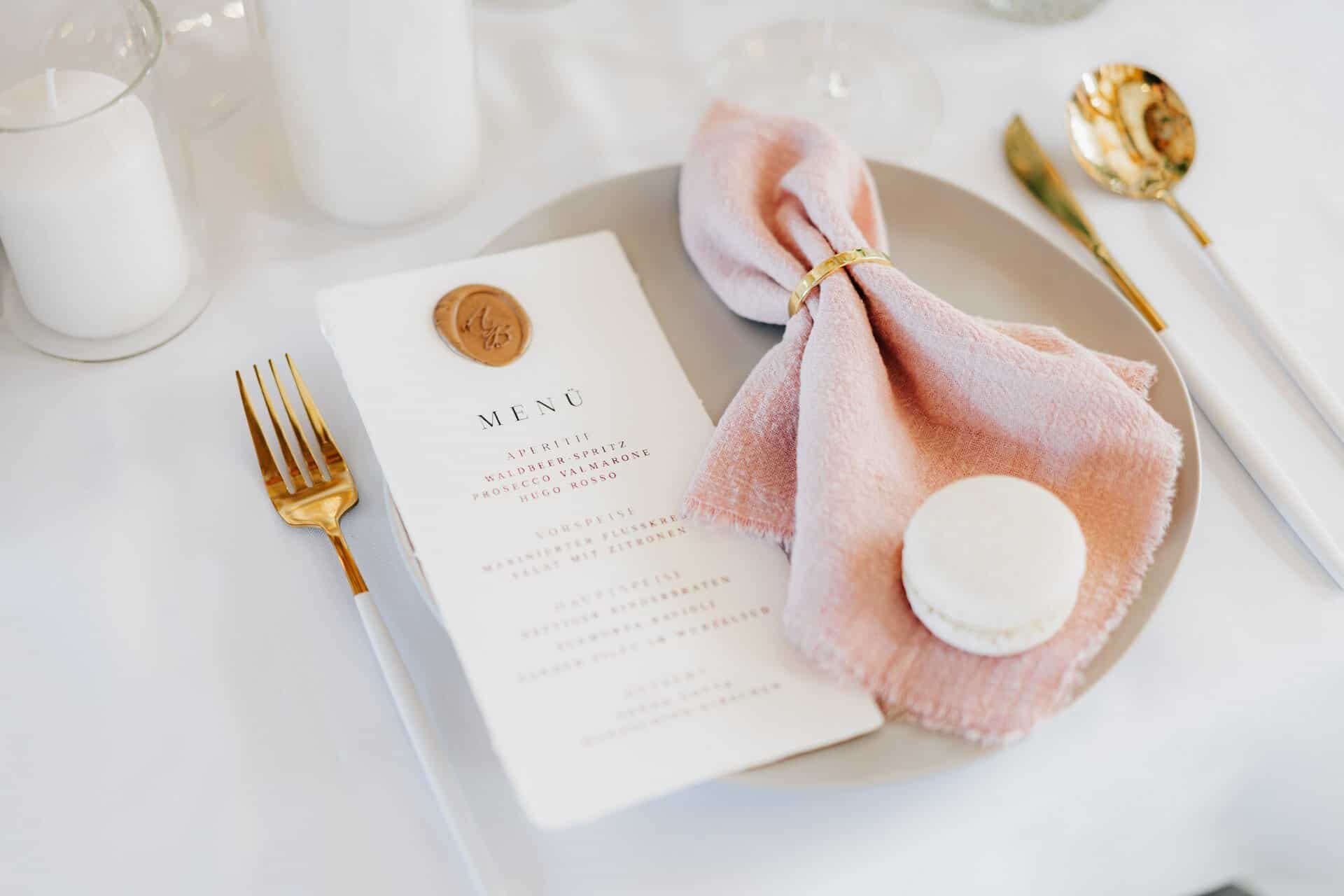 Ein grauer Teller mit einer rosafarbenen Stoffserviette, die von einem goldenen Ring gehalten wird, ein weißer Macaron, eine mit Wachs versiegelte Speisekarte und goldenes Besteck liegen auf einem weißen Tischtuch, auf dem Kerzen brennen - perfekt, um von einem Hochzeitsfotografen Göppingen festgehalten zu werden.