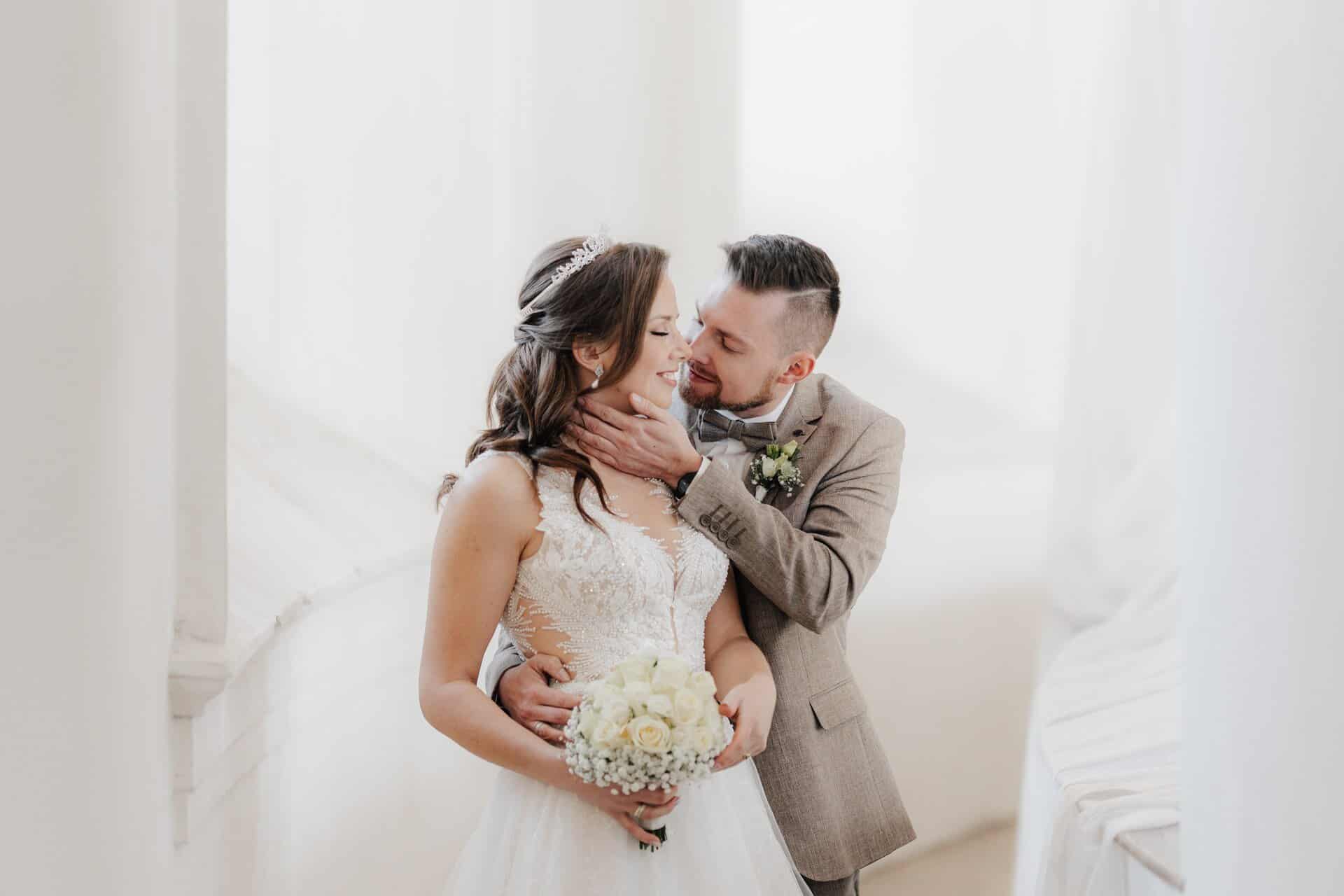 Der Bräutigam hält sanft das Gesicht seiner lächelnden Braut und küsst sie auf die Wange. In einer hellen, luftigen Umgebung fängt der Hochzeitsfotograf Göppingen das Paar eng zusammen ein - die Braut in einem weißen Kleid mit einem Strauß weißer Blumen.