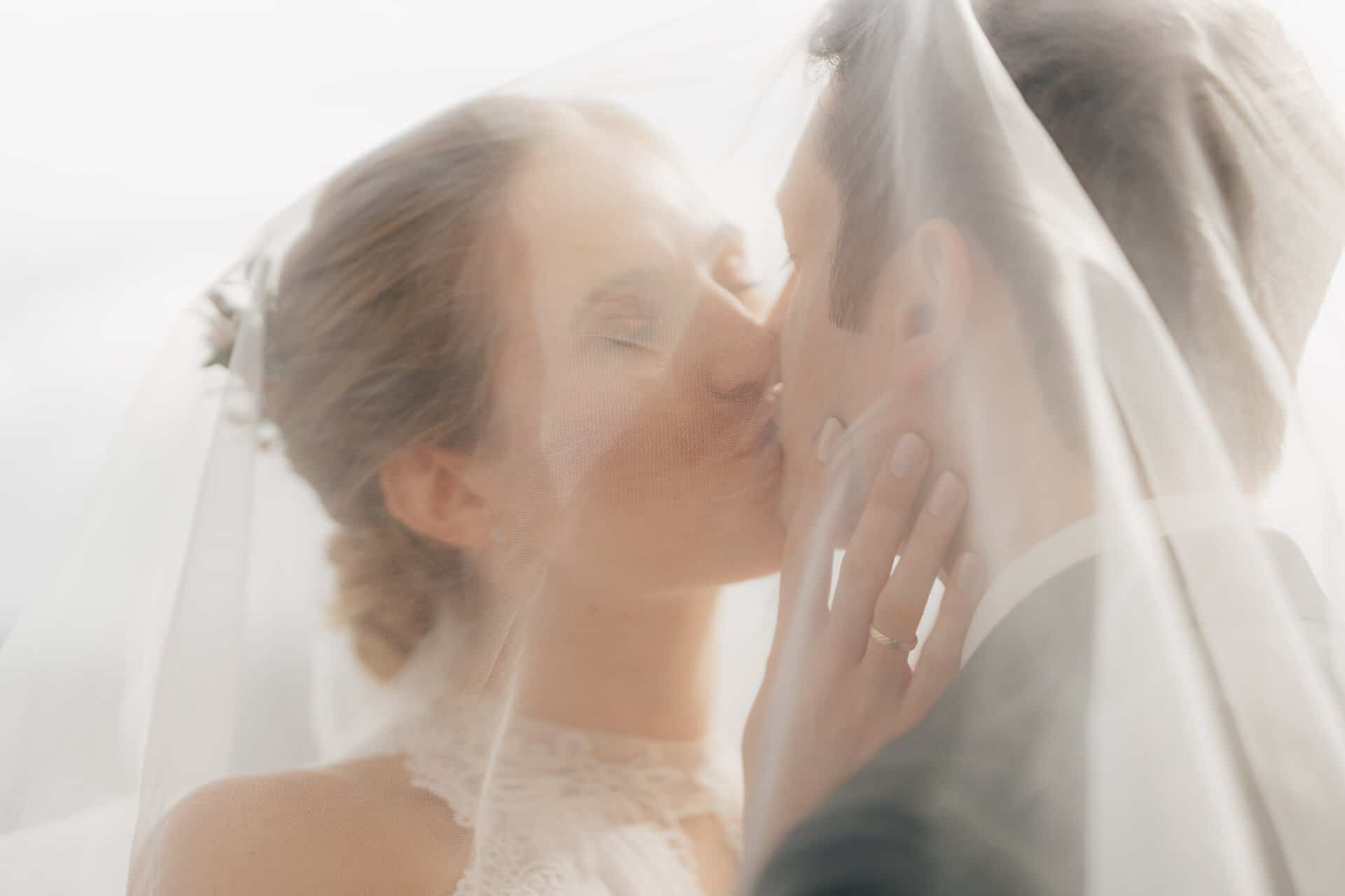 Eine Braut und ein Bräutigam küssen sich zärtlich unter einem durchsichtigen Brautschleier, wobei das weiche Licht ihren intimen Moment hervorhebt. Die Hand der Braut mit dem Ehering liegt auf der Wange des Bräutigams, aufgenommen von Hochzeitsfotograf Göppingen.